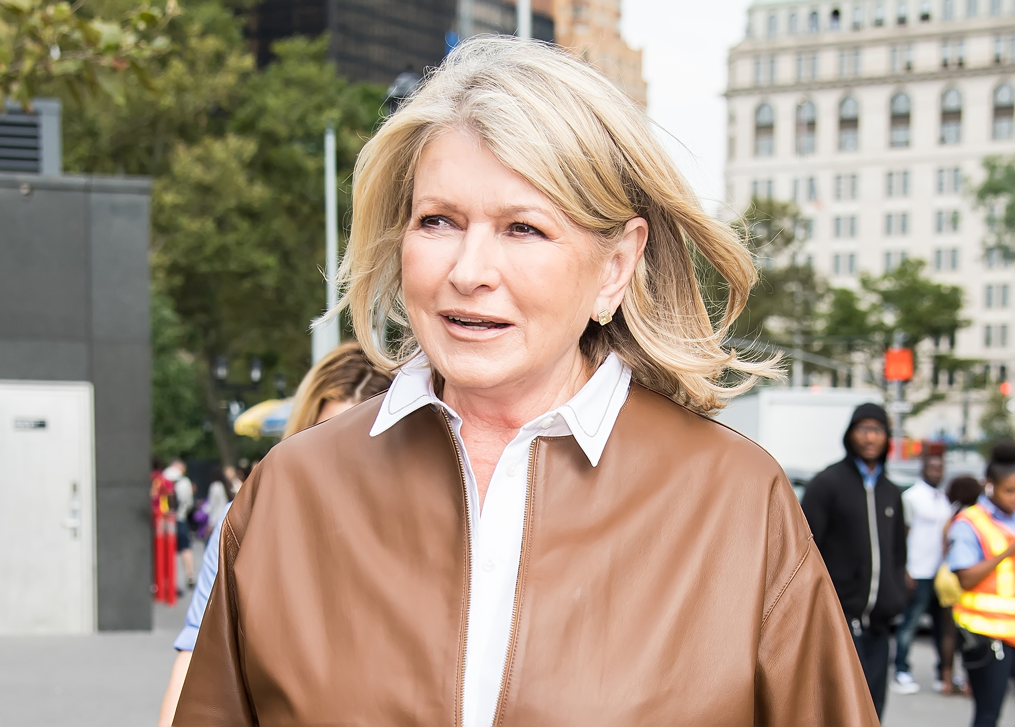 Martha Stewart walks outdoors in a city, wearing a brown jacket over a white top, with a background of buildings and people