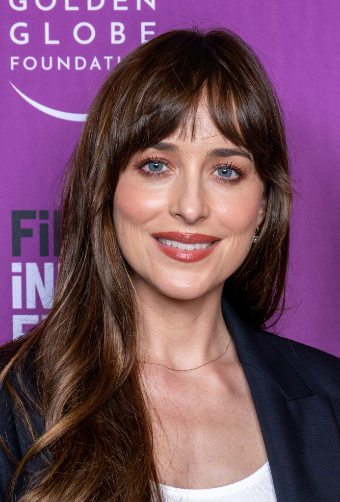 Dakota Johnson at an event, wearing a dark blazer and a white top, smiling at the camera