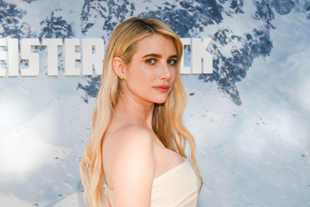 Emma Roberts poses against a snowy mountain backdrop at a red carpet event. She is wearing an elegant, strapless dress