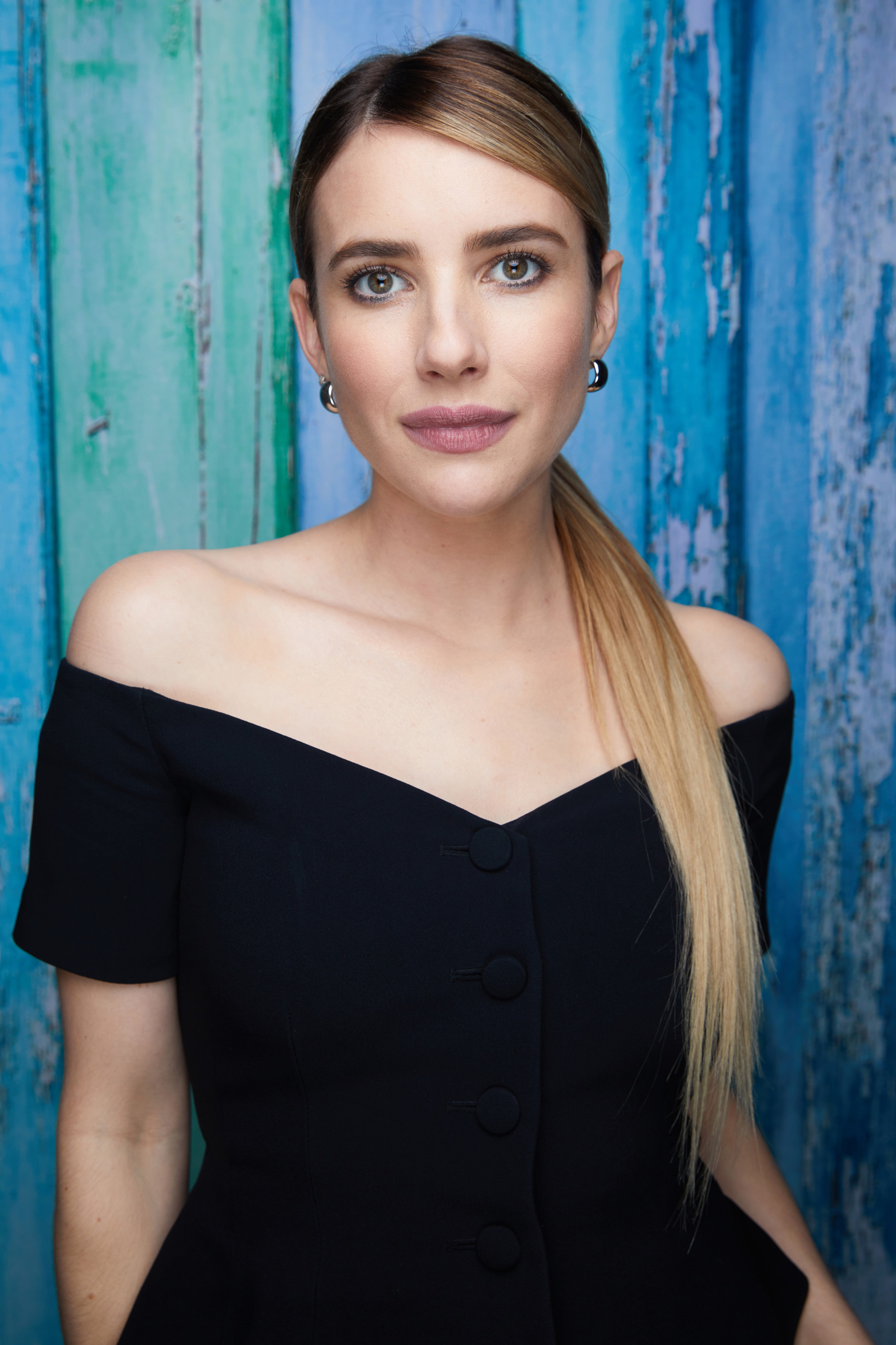 Emma Roberts poses in an off-shoulder black dress against a textured blue and green background