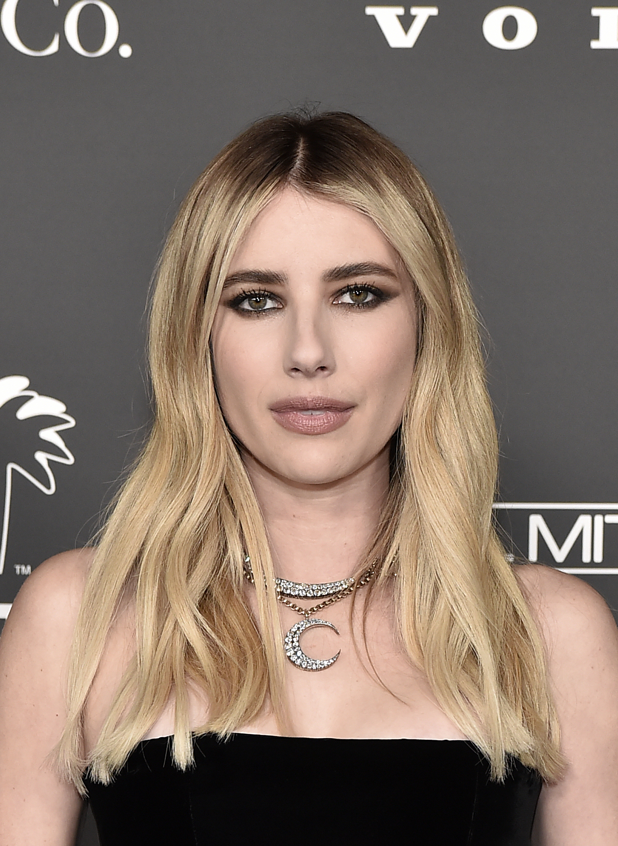 Emma Roberts poses for a photo at a celebrity event, wearing a strapless black dress and layered necklaces against a media backdrop