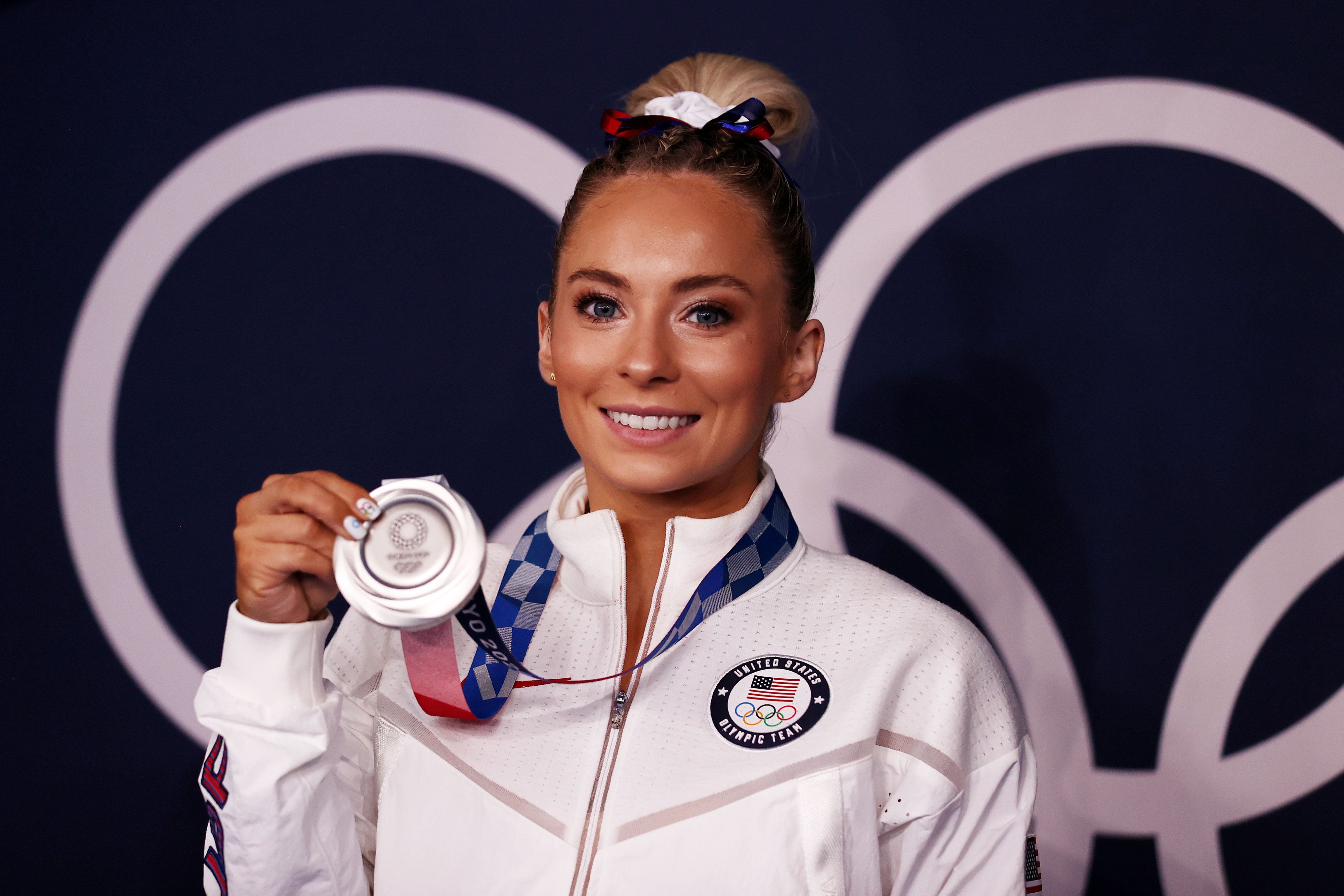 MyKayla Skinner holds her silver Olympic medal while smiling in front of an Olympic rings backdrop. She is wearing a Team USA tracksuit