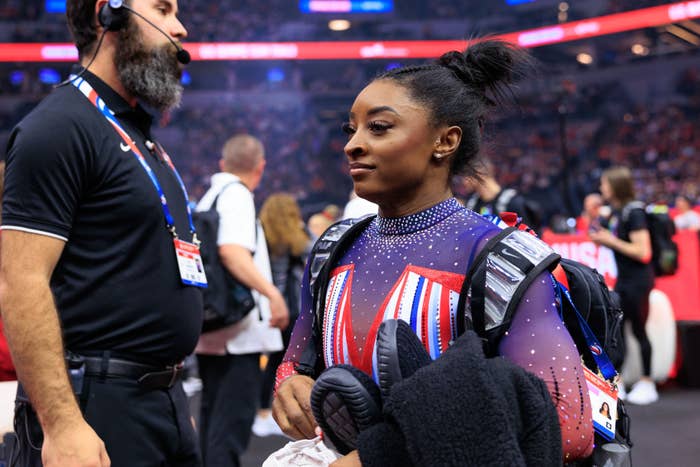 Simone Biles stands in a gymnastics arena wearing a sparkly leotard and holding a jacket, with an event official nearby