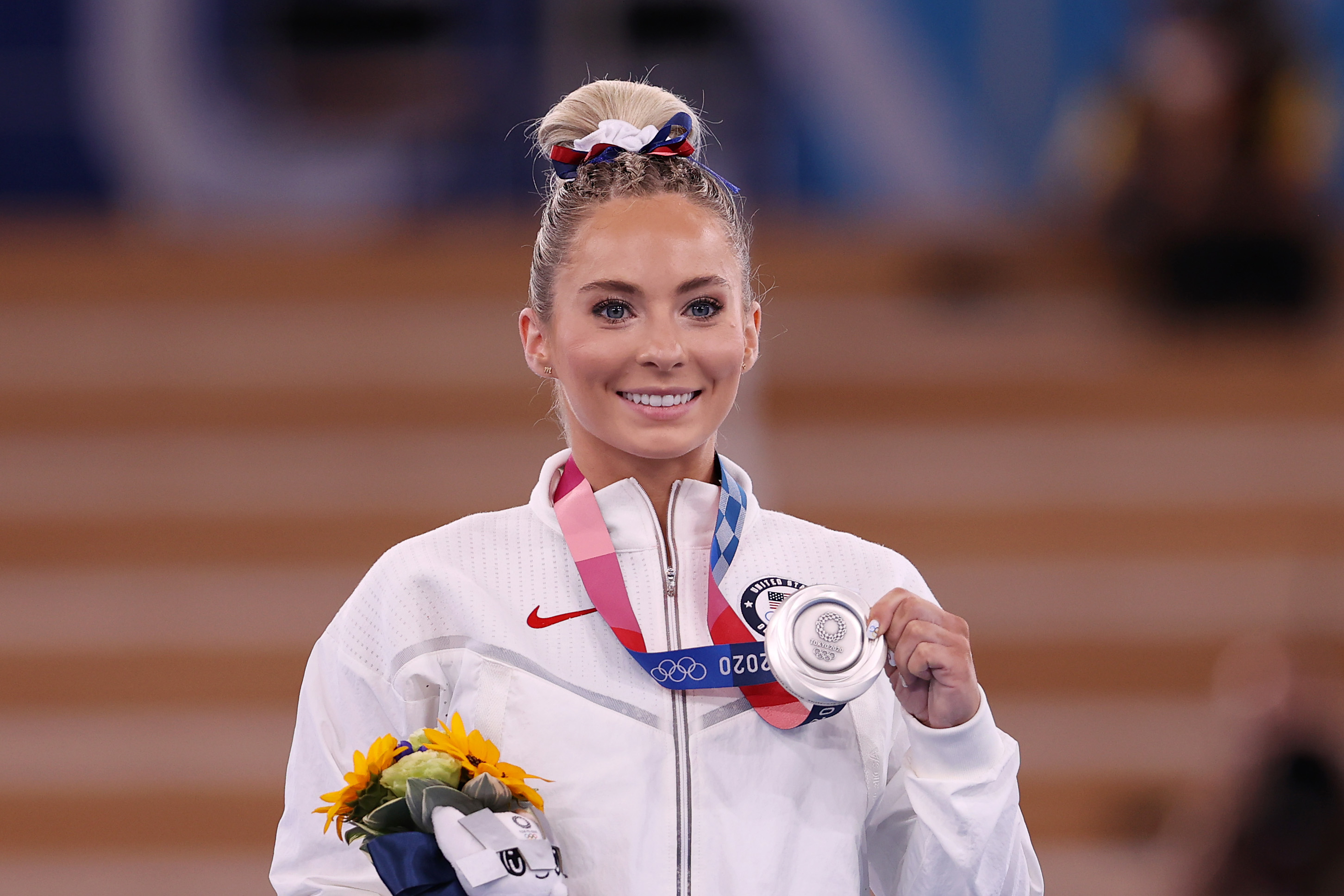 MyKayla Skinner smiles and holds her silver medal on the podium at the Tokyo 2020 Olympics. She is wearing a white athletic jacket and holding a bouquet of flowers