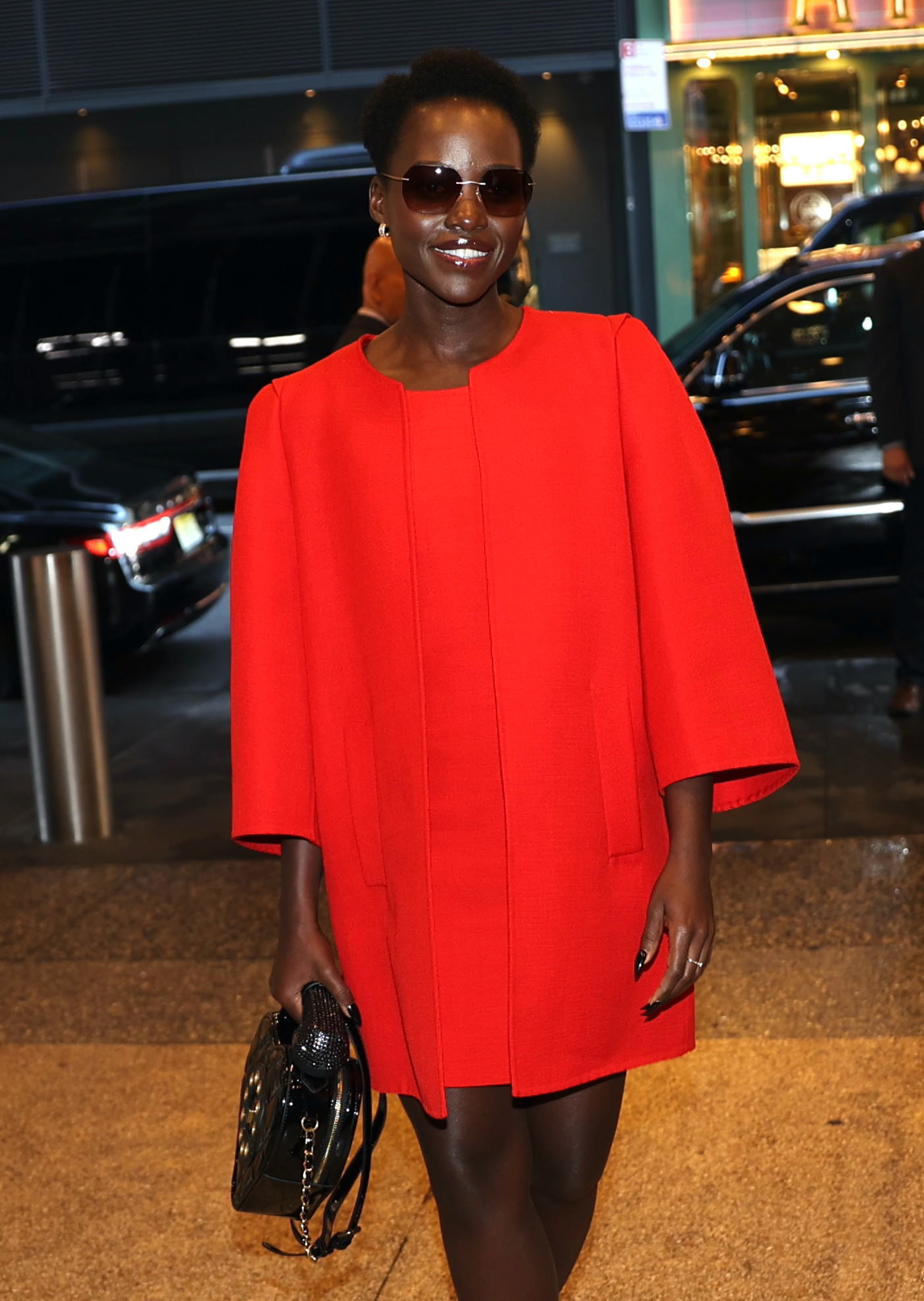 Lupita Nyong&#x27;o is walking in a city at night wearing sunglasses and a stylish knee-length cape dress. She is holding a black handbag and smiling