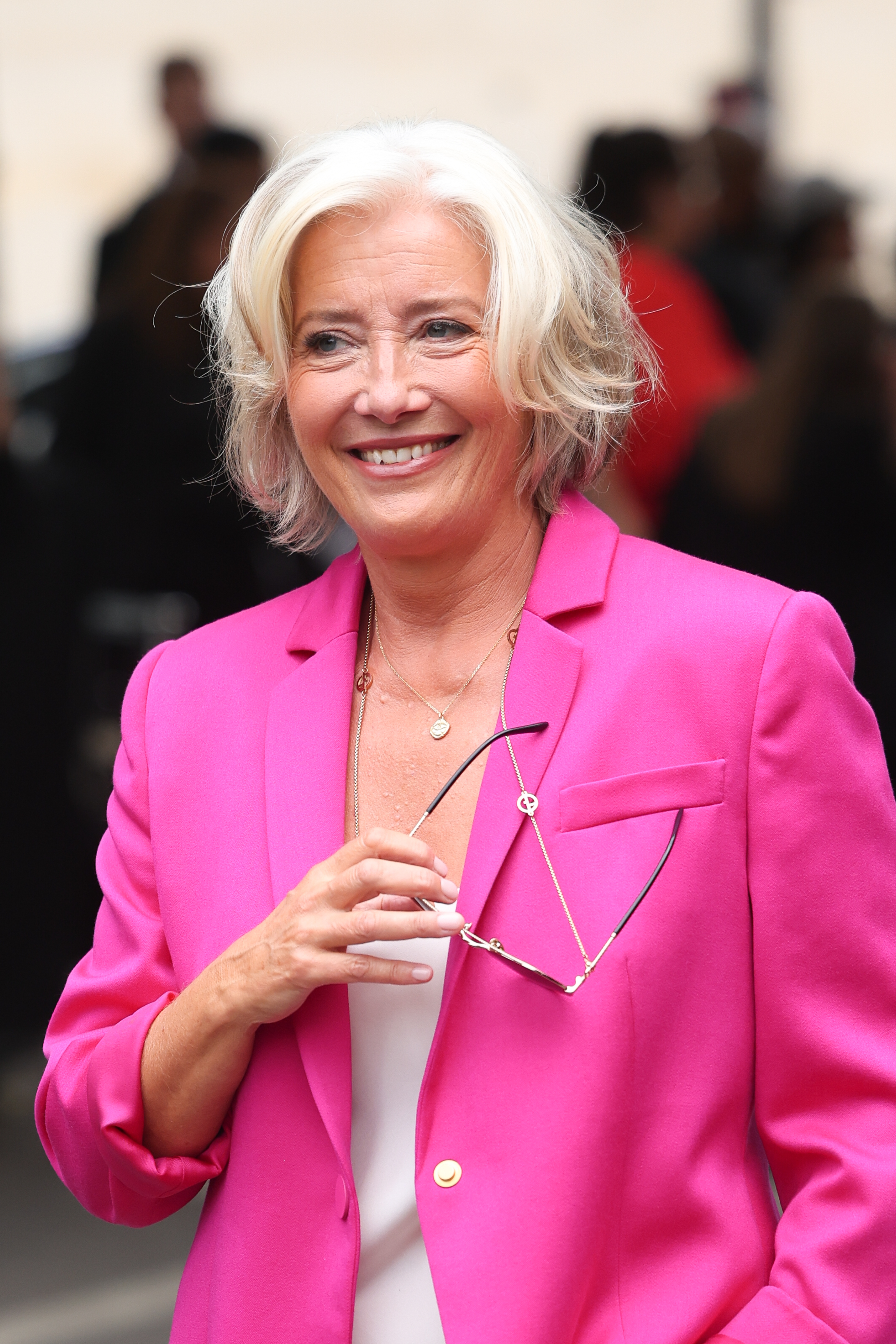 Emma Thompson smiles, holding eyeglasses, wearing a stylish blazer over a simple blouse