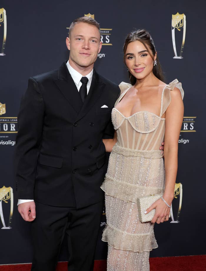 Christian McCaffrey and Olivia Culpo pose together on a red carpet. Christian wears a double-breasted suit, and Olivia wears a sheer, tiered gown