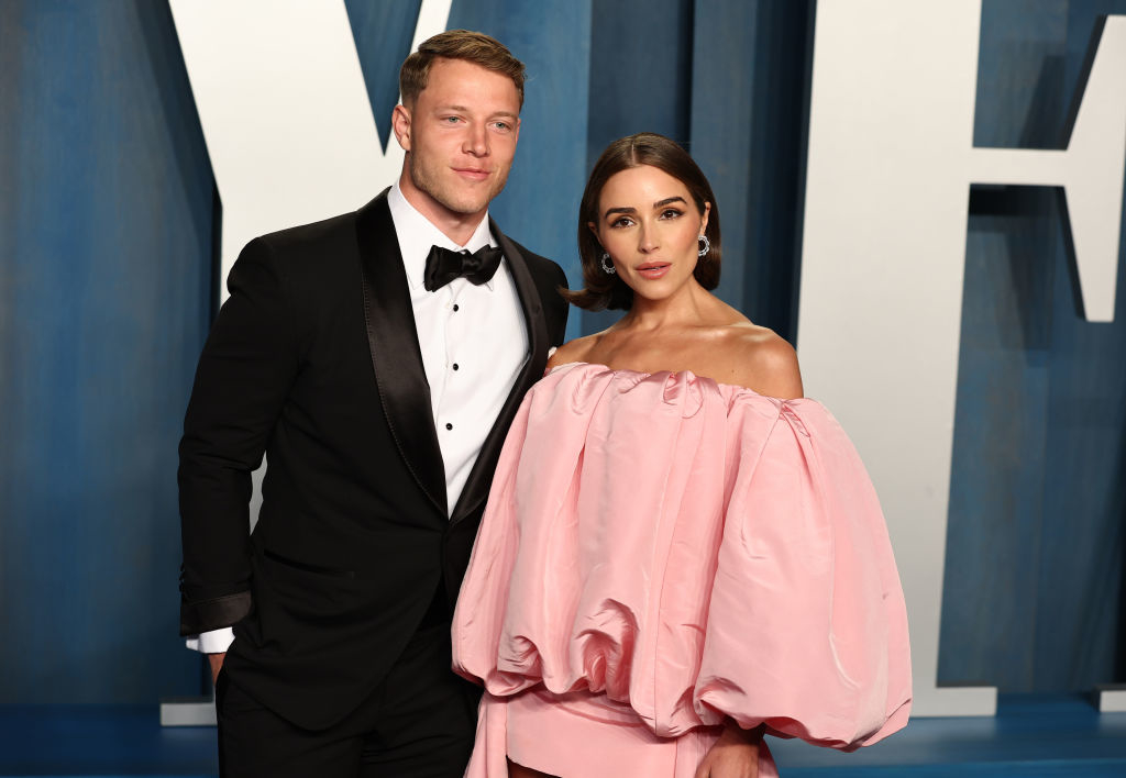 Christian McCaffrey and Olivia Culpo pose together at a formal event. Christian wears a black tuxedo, while Olivia wears an off-the-shoulder pink gown