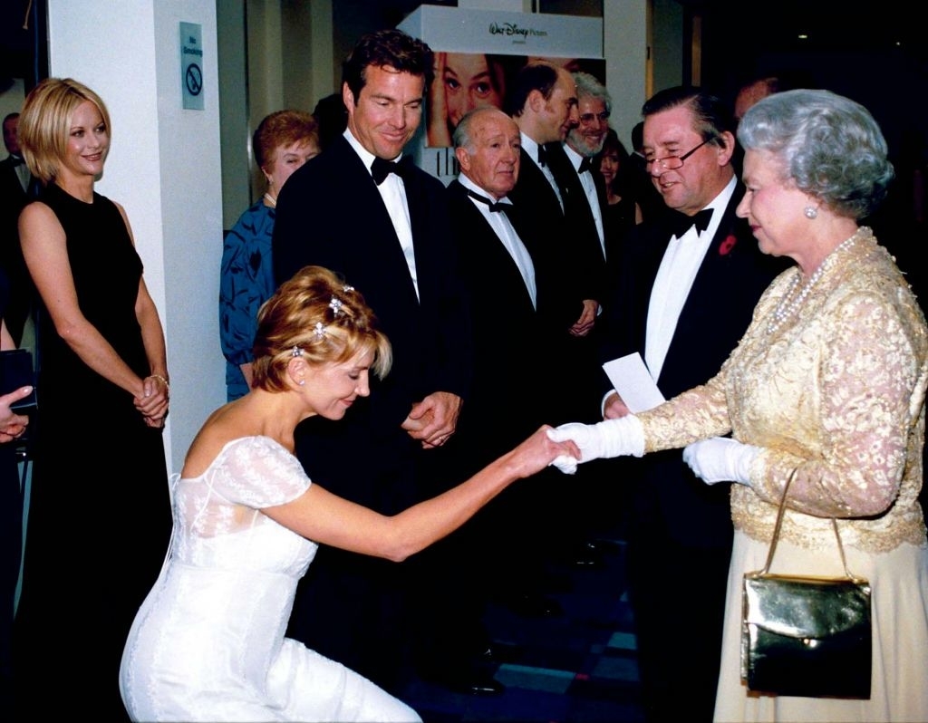 Helen Mirren in a white dress curtsies to Queen Elizabeth II, who is wearing a golden outfit and gloves, at an event. Several people are in the background