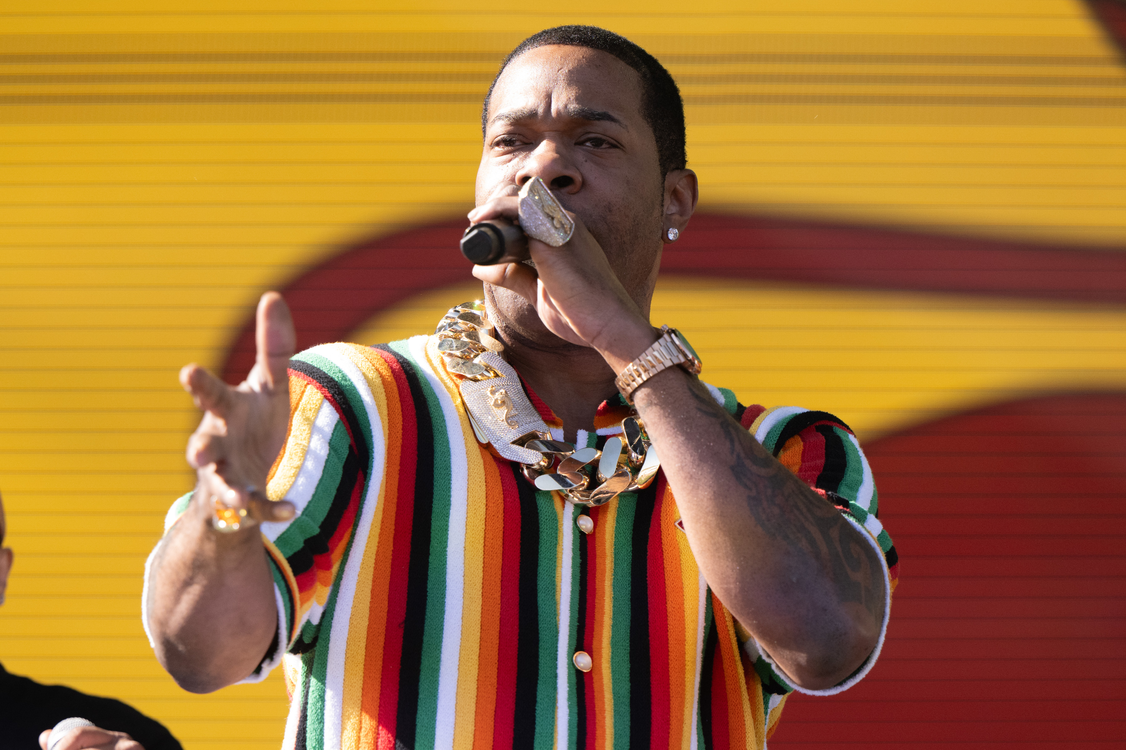 Busta Rhymes performs on stage, wearing a colorful striped shirt and a large gold chain necklace