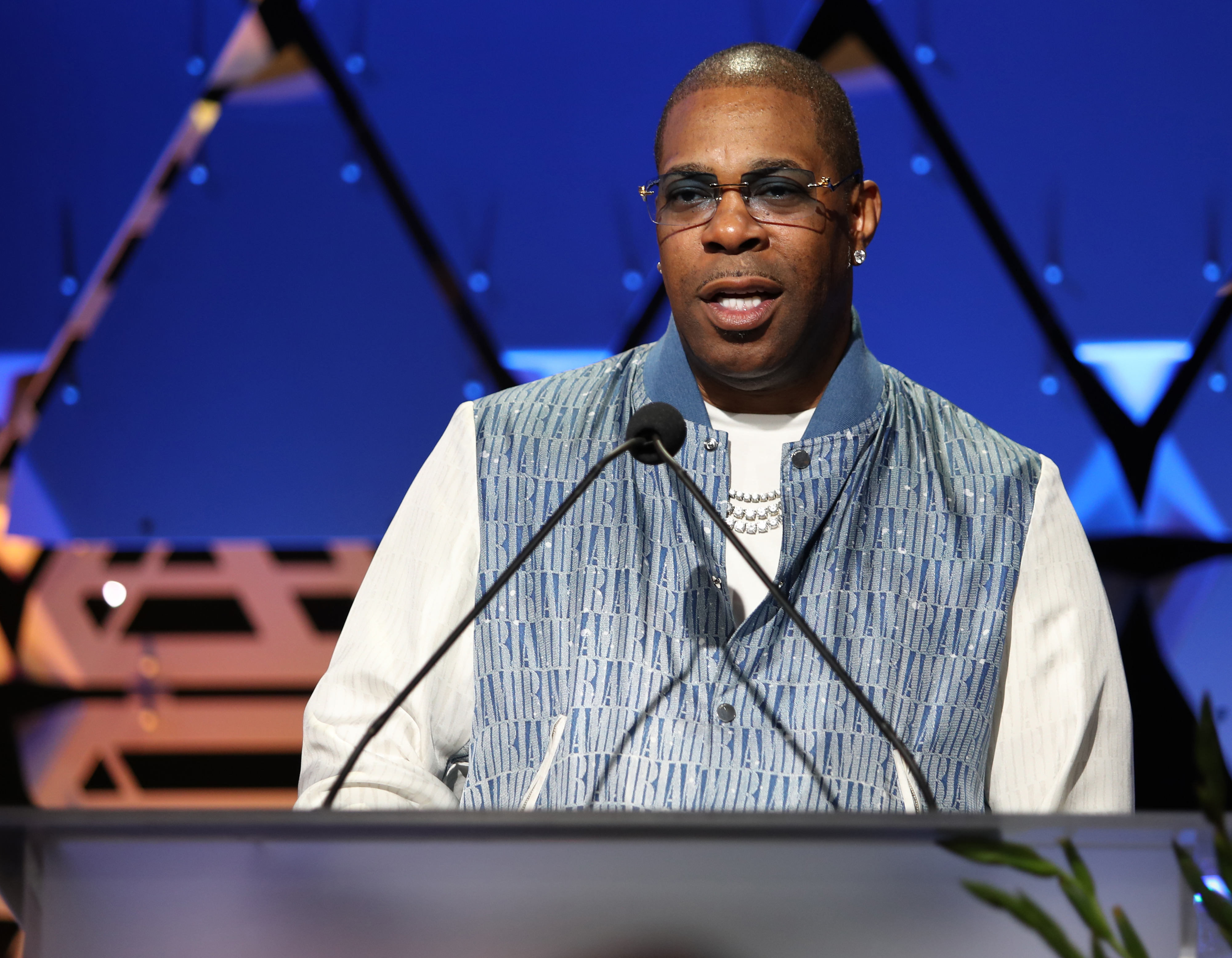 Busta Rhymes speaks at a podium, dressed in a white shirt and patterned vest, with sunglasses and jewelry