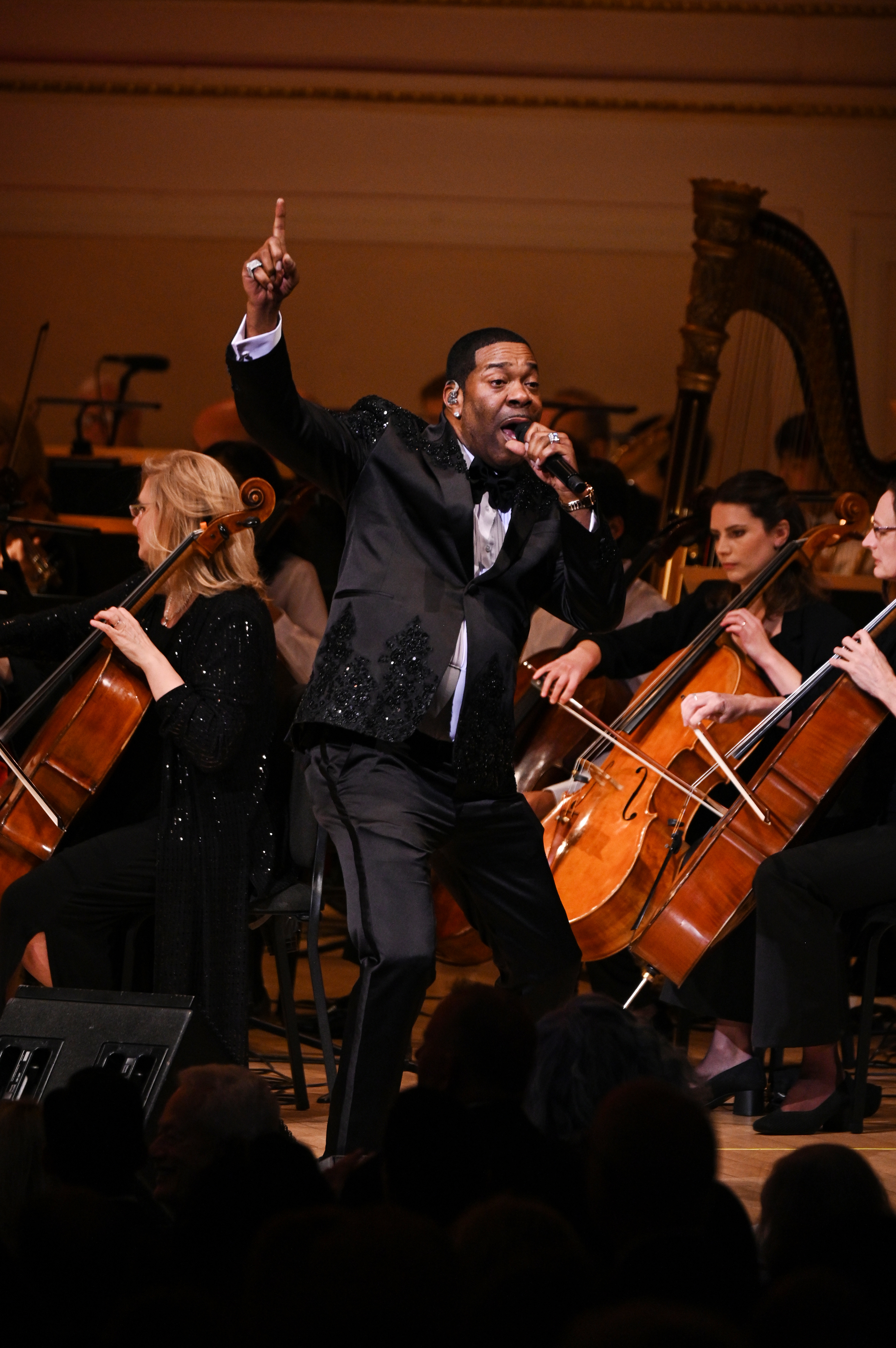 Busta Rhymes performing in front of an orchestra, wearing a formal black suit with shimmering accents, passionately singing into a microphone