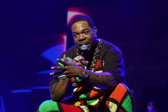 Busta Rhymes performs on stage, wearing a colorful knit outfit and large gold chains, while holding a microphone and gesturing expressively with his hands