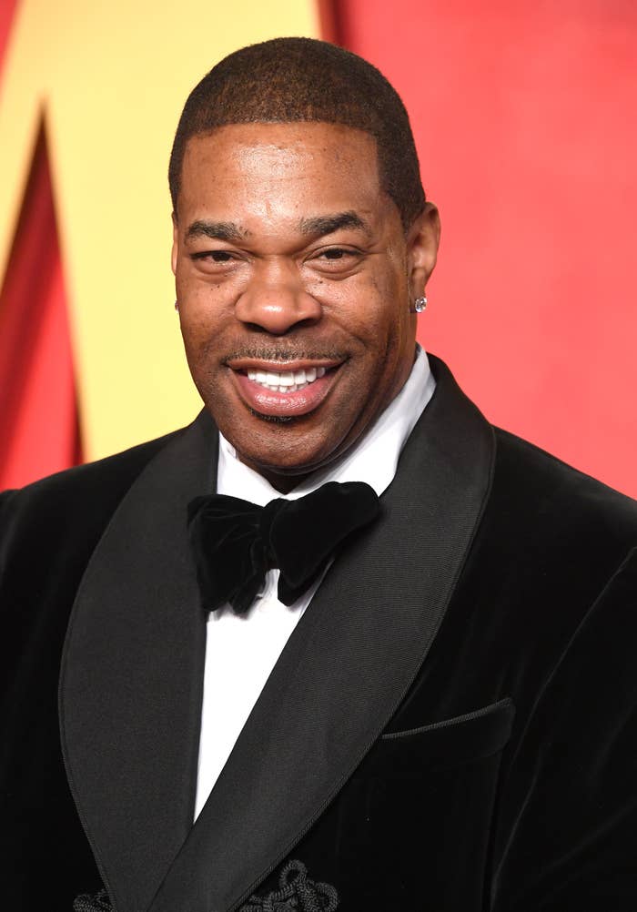 Busta Rhymes at a red carpet event, wearing a velvet tuxedo jacket with a bow tie, smiling at the camera