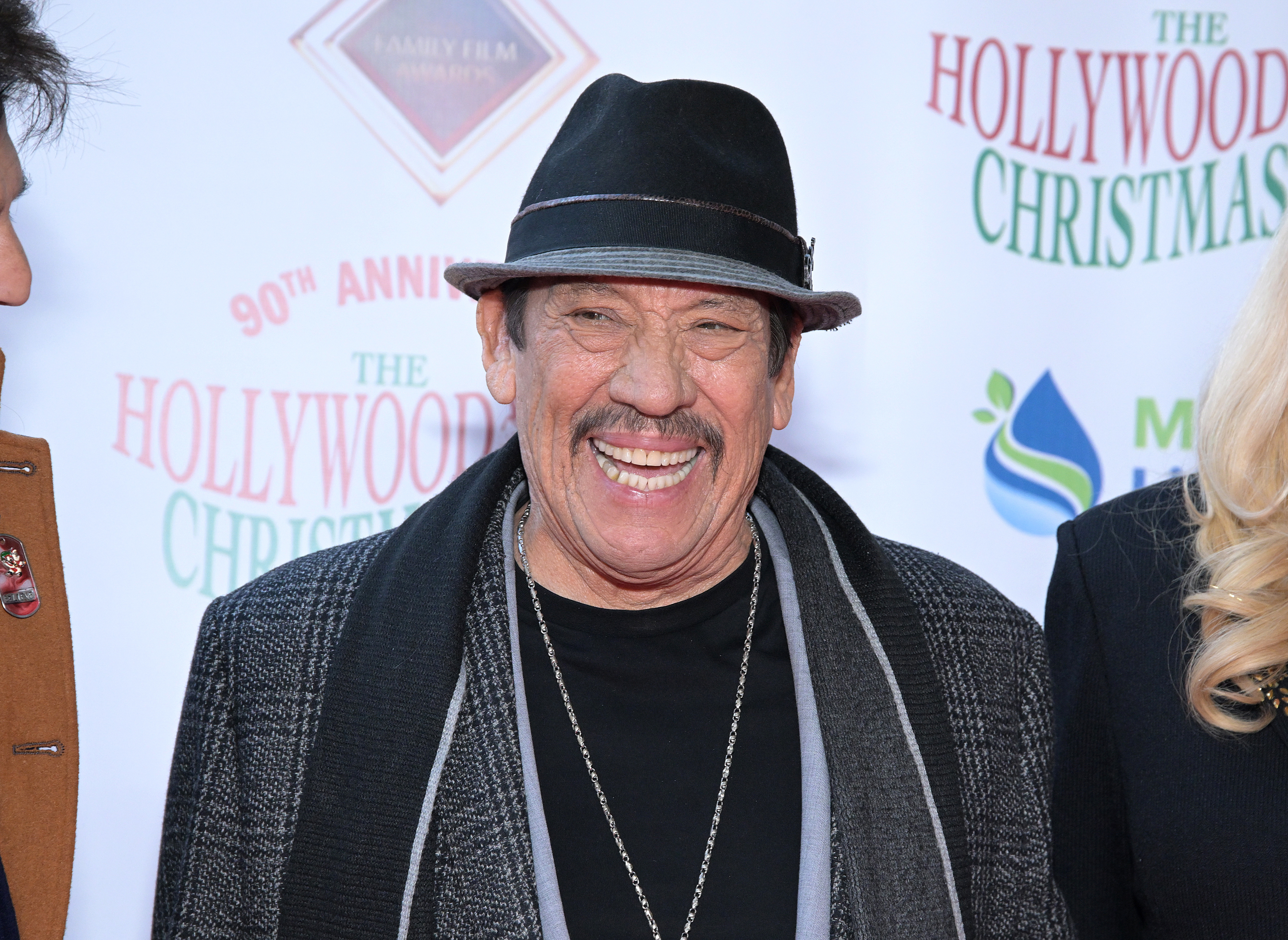 Danny Trejo wearing a hat and a long coat smiles at the 90th Annual Hollywood Christmas Parade event