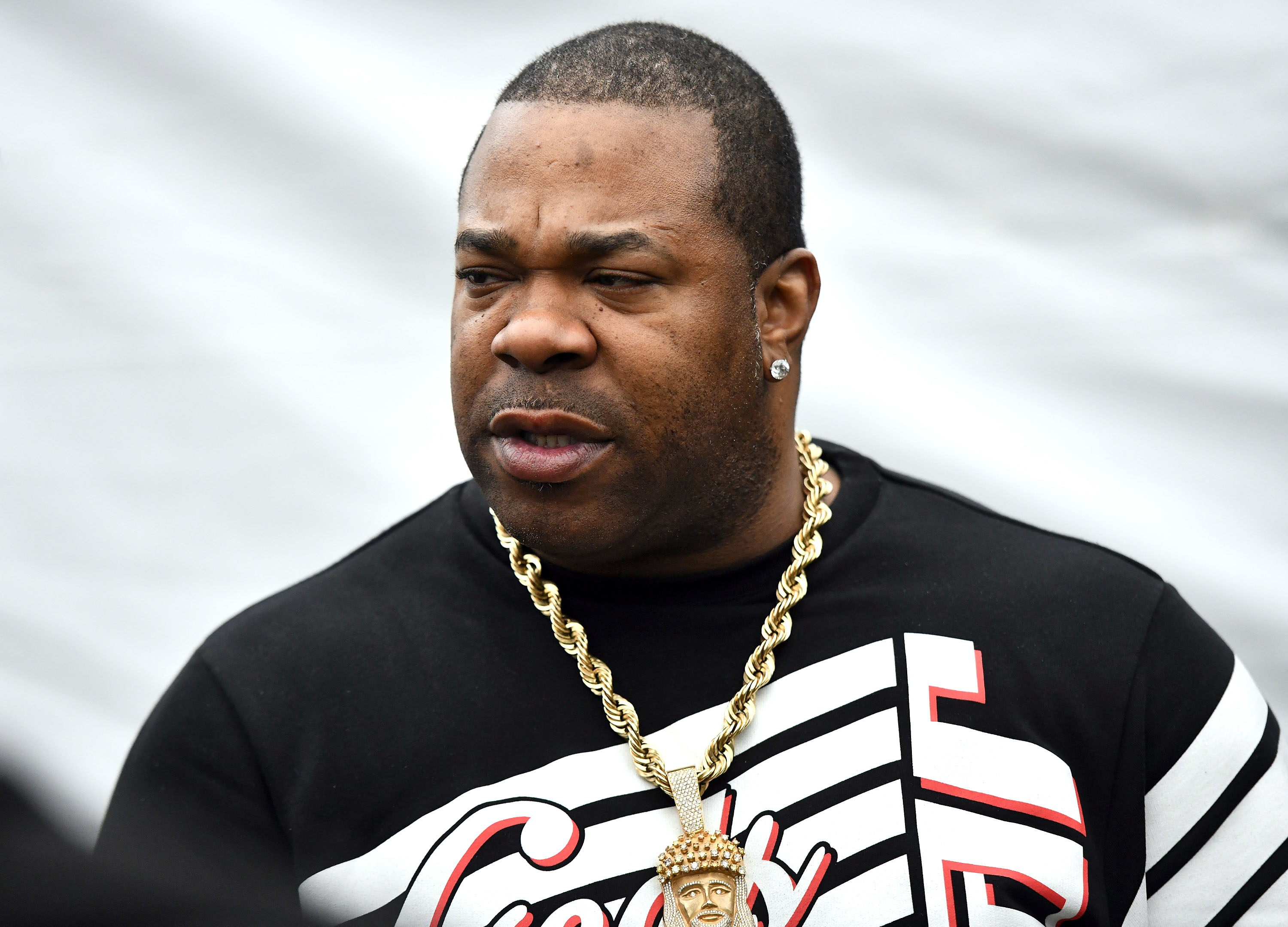 Busta Rhymes wearing a black and white graphic shirt and a gold chain necklace, looking to the side with a neutral expression