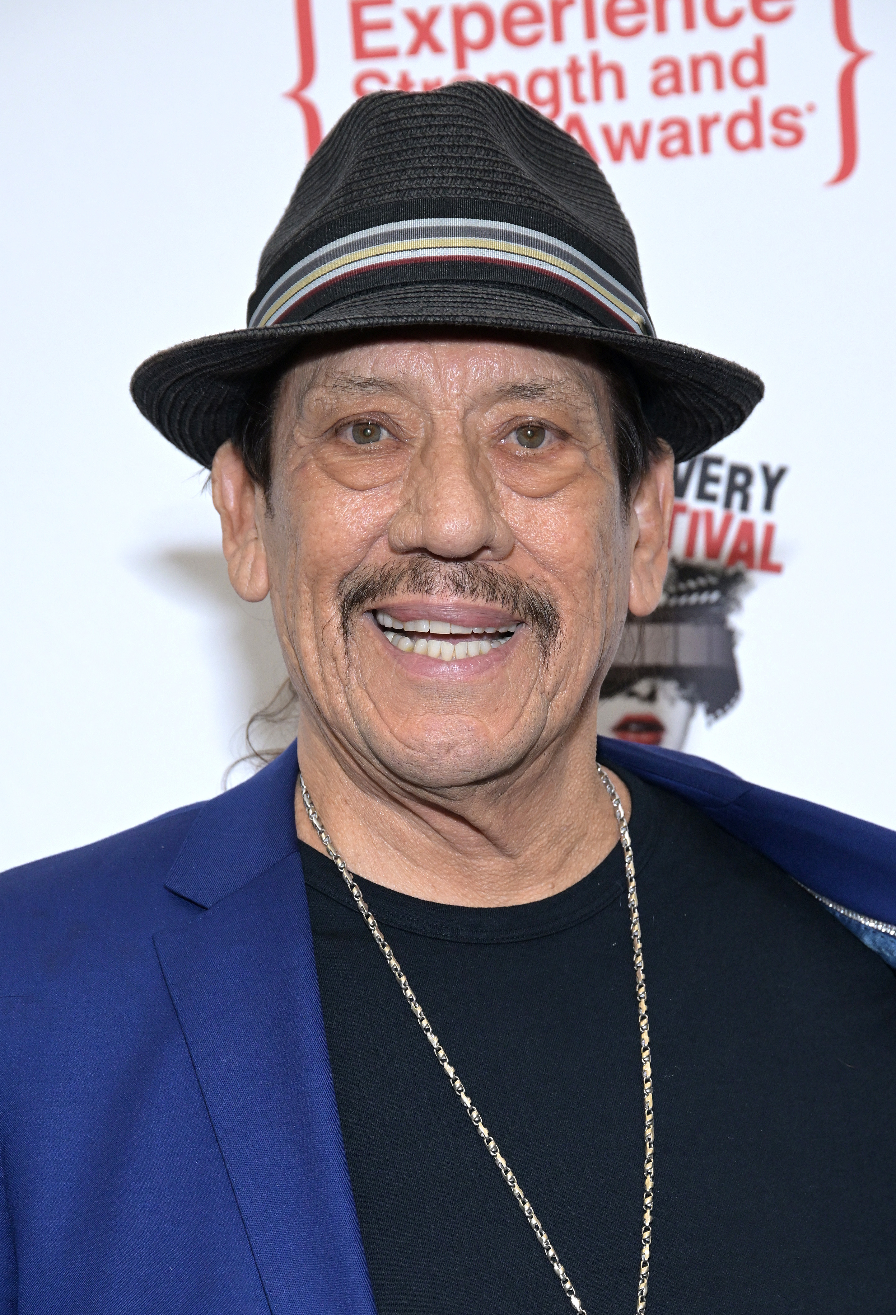 Danny Trejo wearing a hat, black shirt, and blue blazer at an event