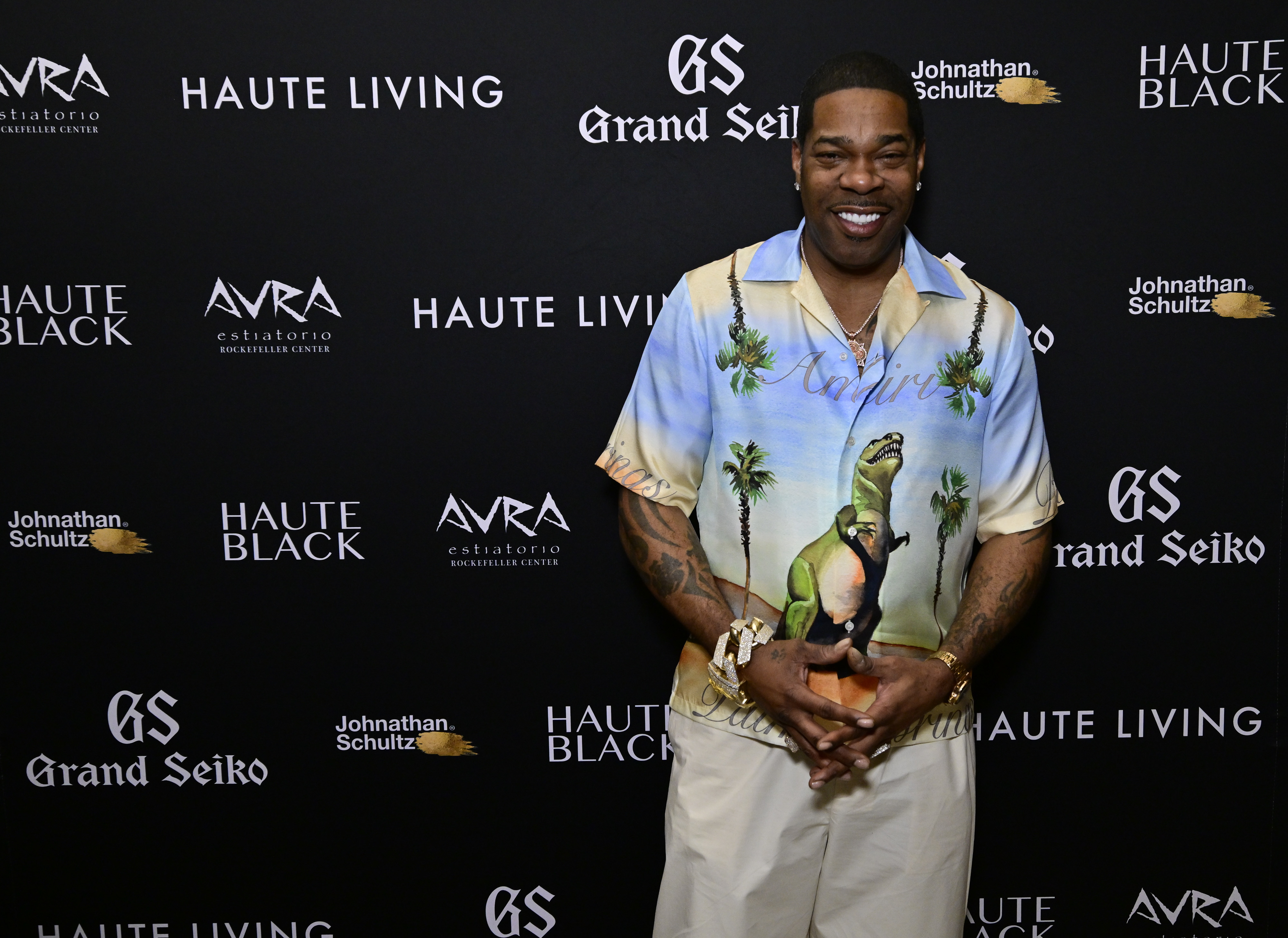 Busta Rhymes smiles on the red carpet, wearing a printed short-sleeved shirt with palm trees and white pants, against a Haute Living backdrop