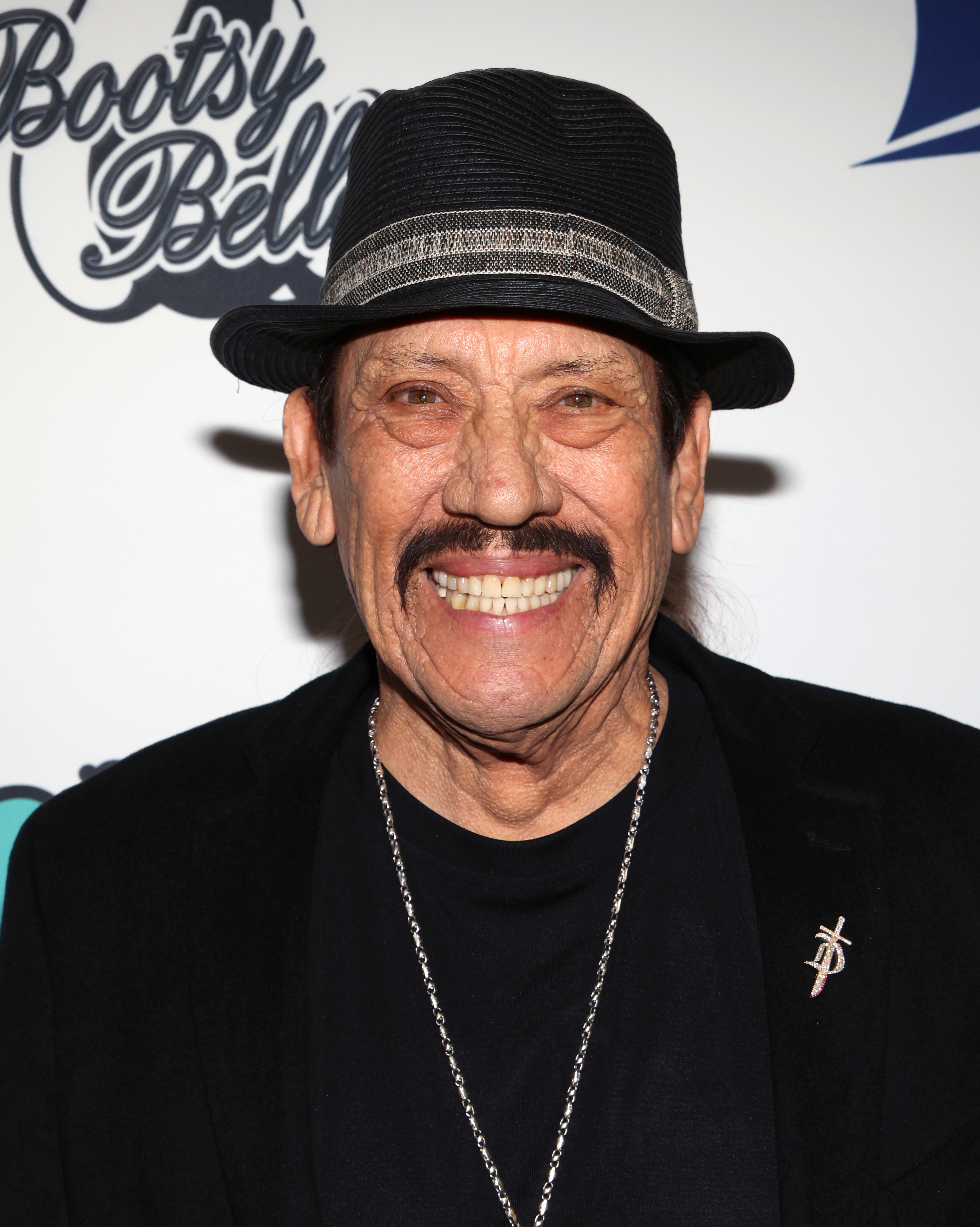 Danny Trejo smiles at an event, wearing a black hat, black blazer, and a silver necklace with a cross pendant