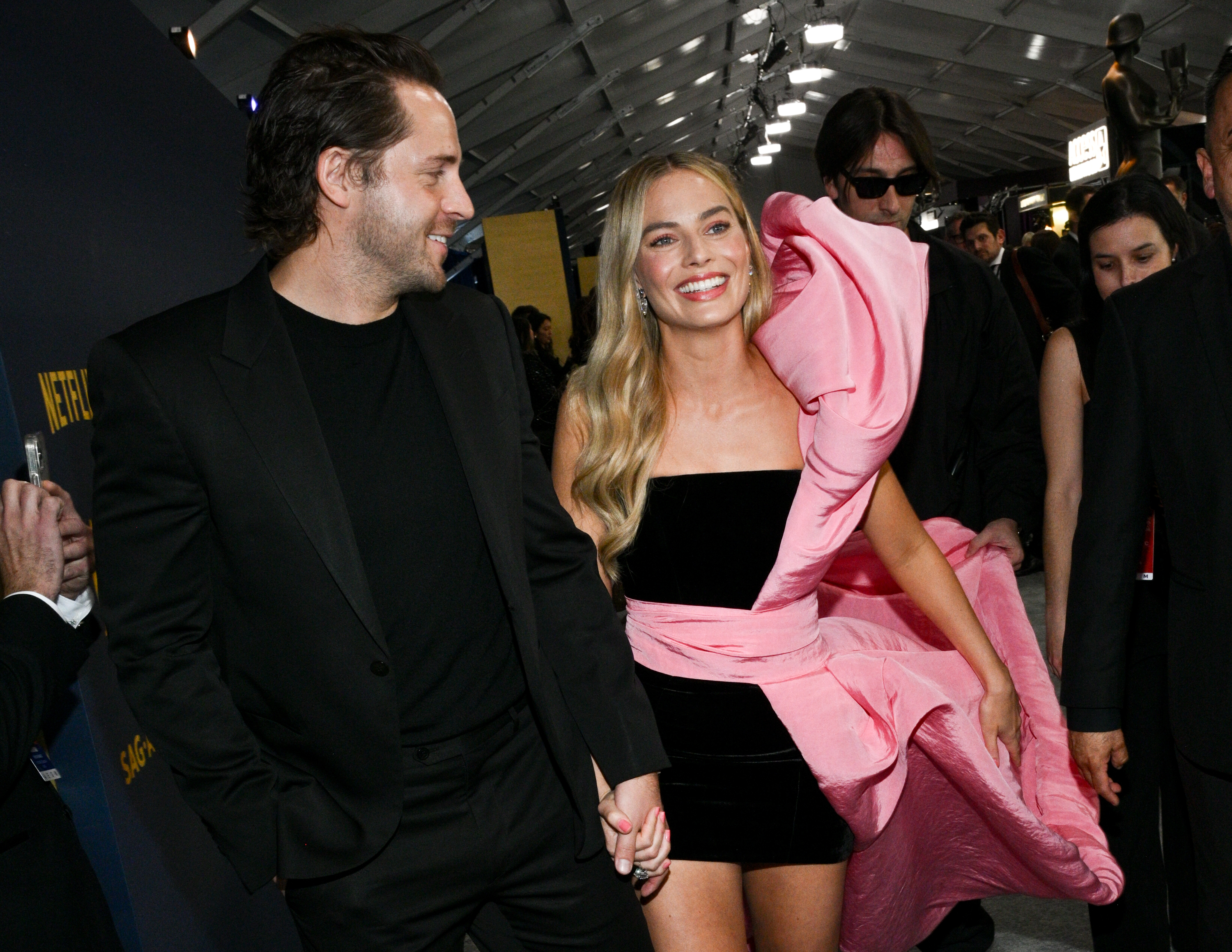 Margot Robbie, in a strapless black dress with a large pink ruffle, holds hands with a man in a black suit at a formal event