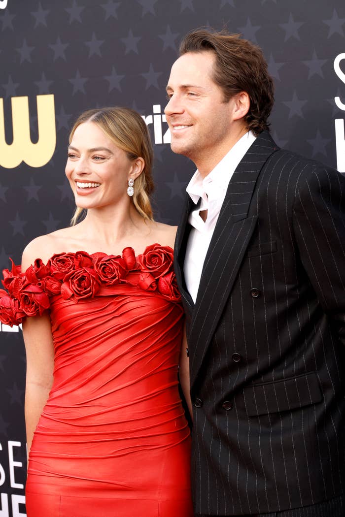 Margot Robbie and Tom Ackerley on a red carpet. Margot wears an off-the-shoulder dress with floral accents, while Tom wears a pinstriped suit. Both are smiling