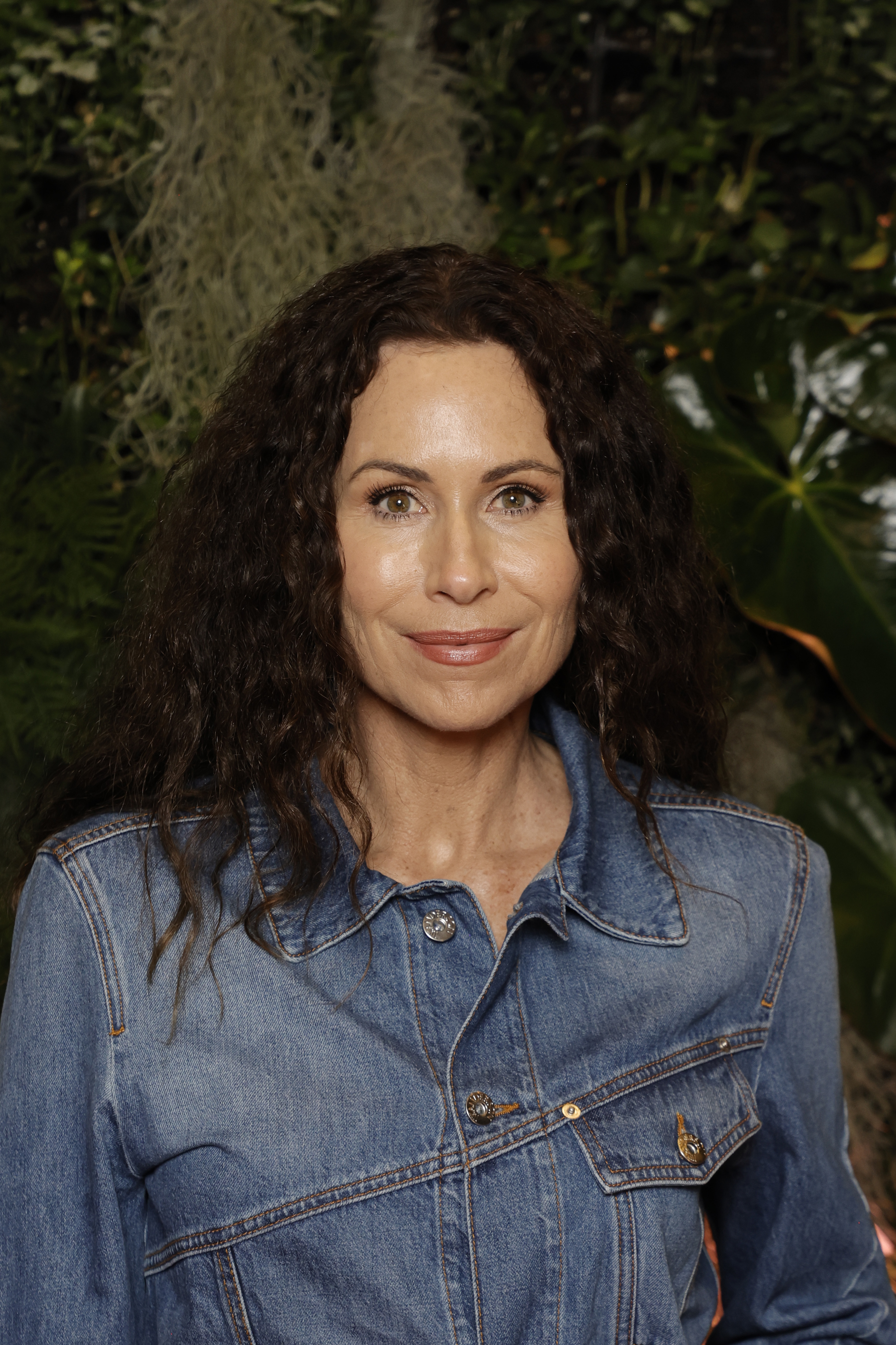 Minnie Driver smiles, wearing a denim jacket, standing in front of a leafy background