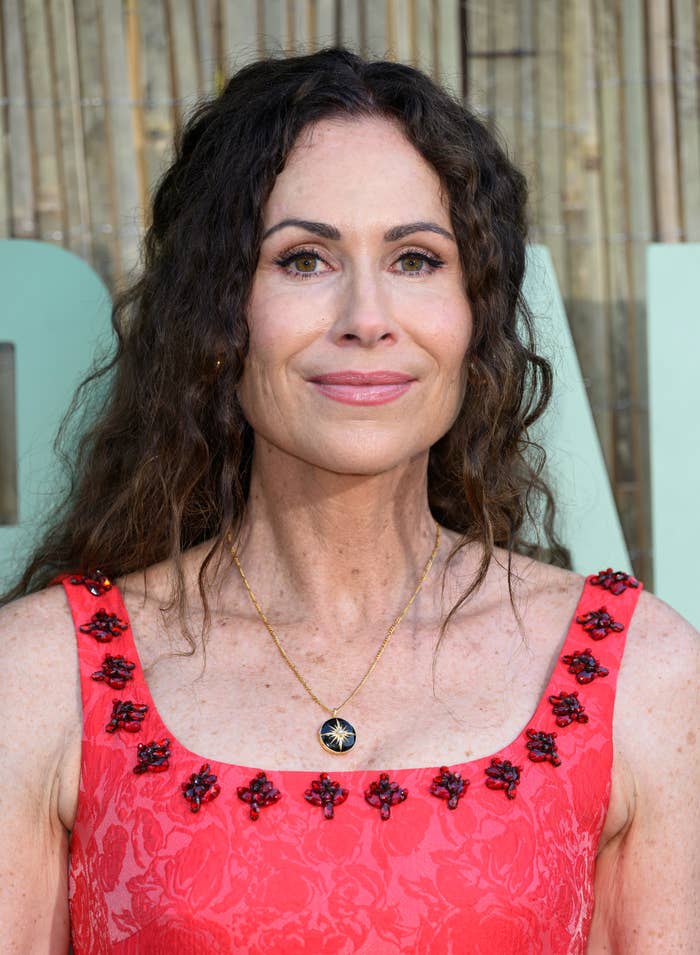 Minnie Driver is wearing a sleeveless dress with floral details and a pendant necklace while smiling at an event