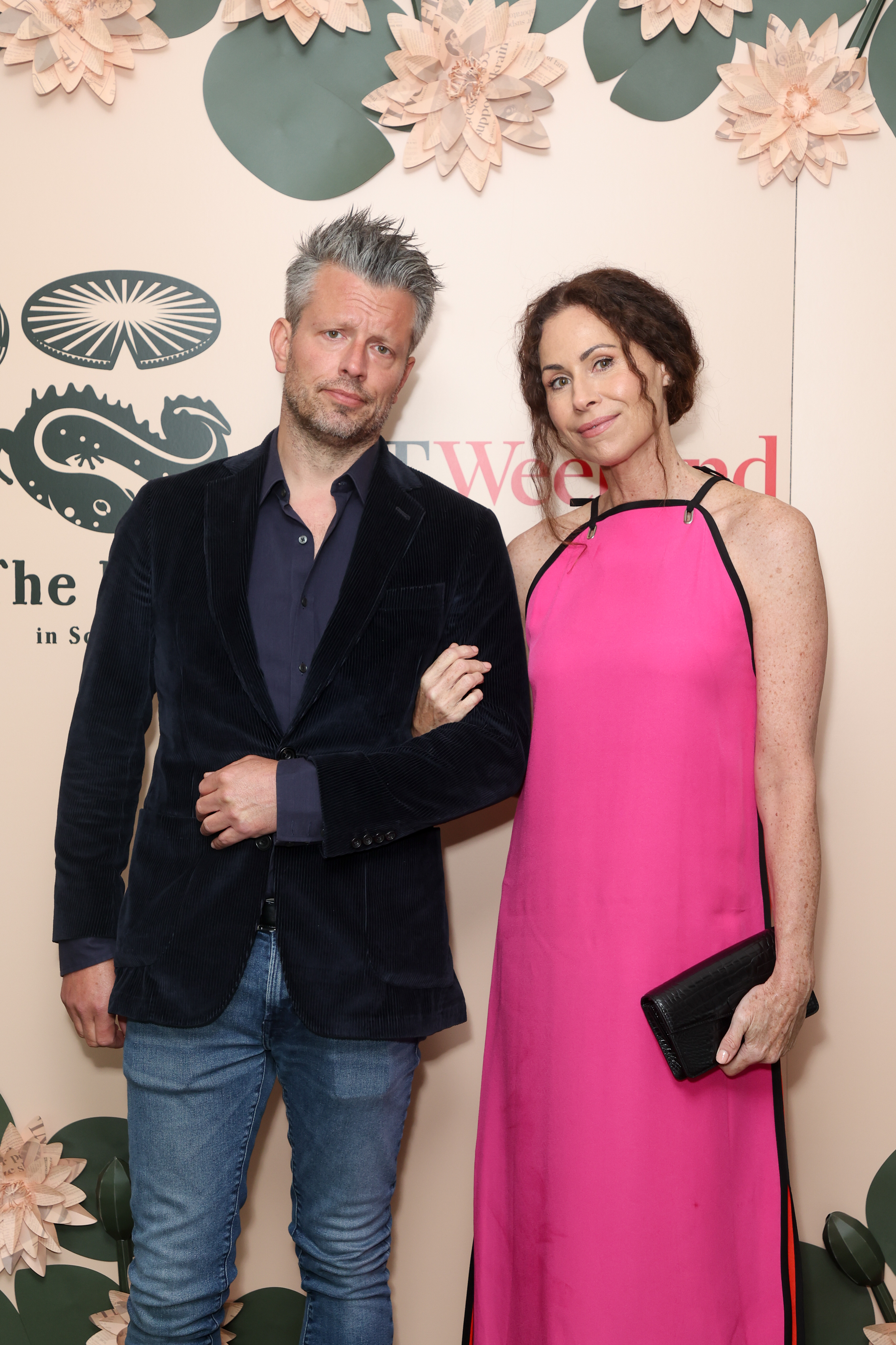Addison O&#x27;Dea and Minnie Driver standing together. The man wears a dark blazer over a shirt and blue jeans. The woman wears a sleeveless pink dress, holding a black clutch