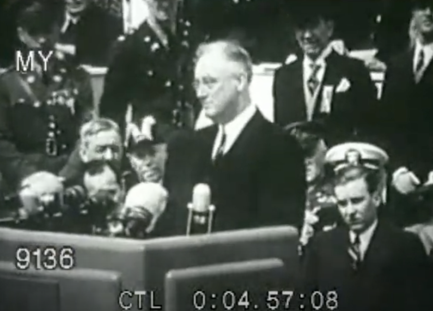 Franklin D. Roosevelt delivers a speech at a historical event. He stands at a podium, with several military and government officials seated behind him