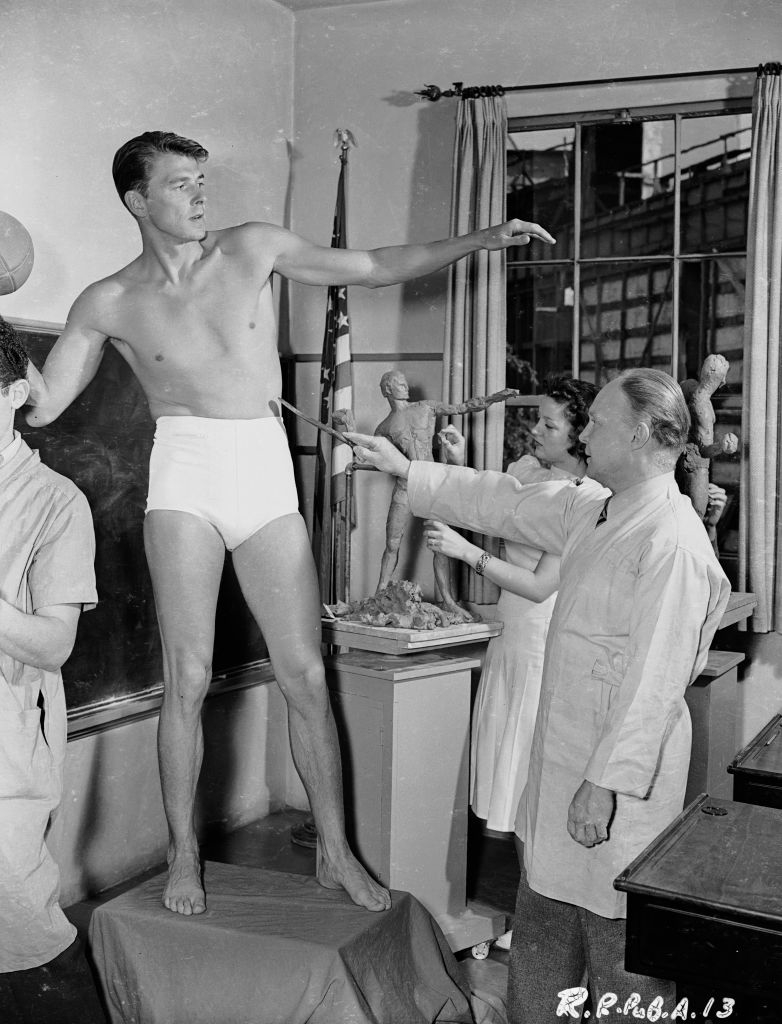 A shirtless young man in shorts stands on a pedestal while others, including an older man in a lab coat, examine and measure him in a classroom setting