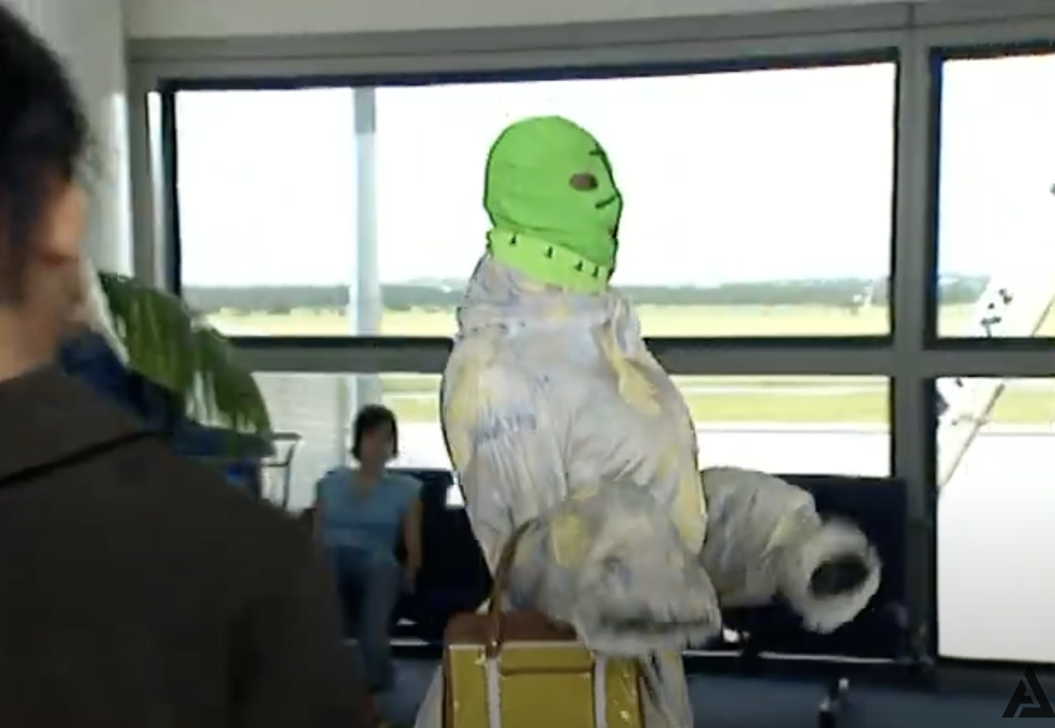 Unidentified person in a green full-face mask and bulky patterned outfit, carrying a tan handbag, standing in what appears to be an airport lounge