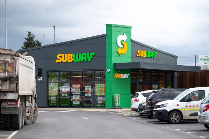 Subway restaurant exterior with several parked vehicles in the lot, including a large truck and white vans. Bright signage visible on the building