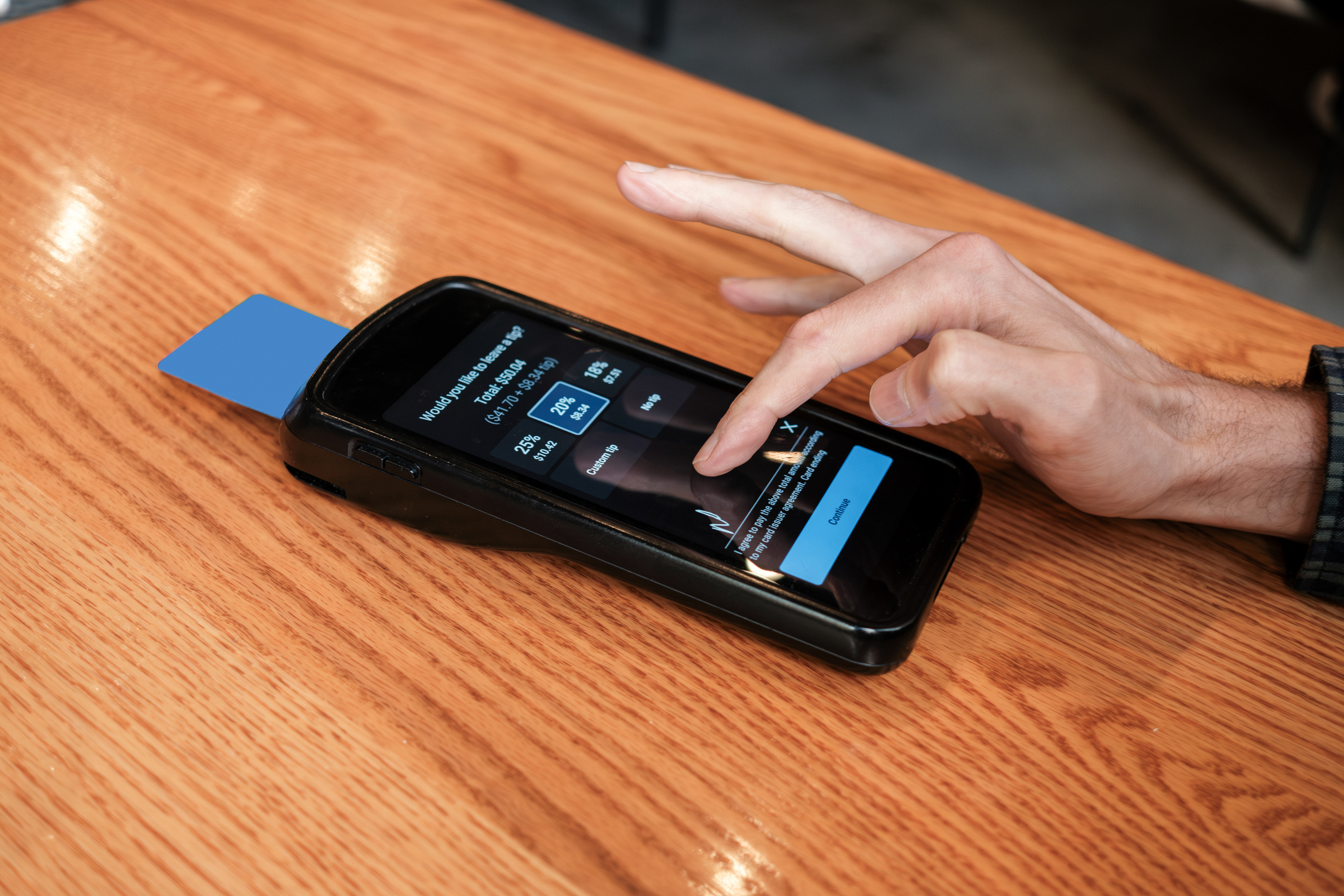 A hand taps a payment terminal screen on a wooden table, where a credit card is inserted. The screen displays options for tipping