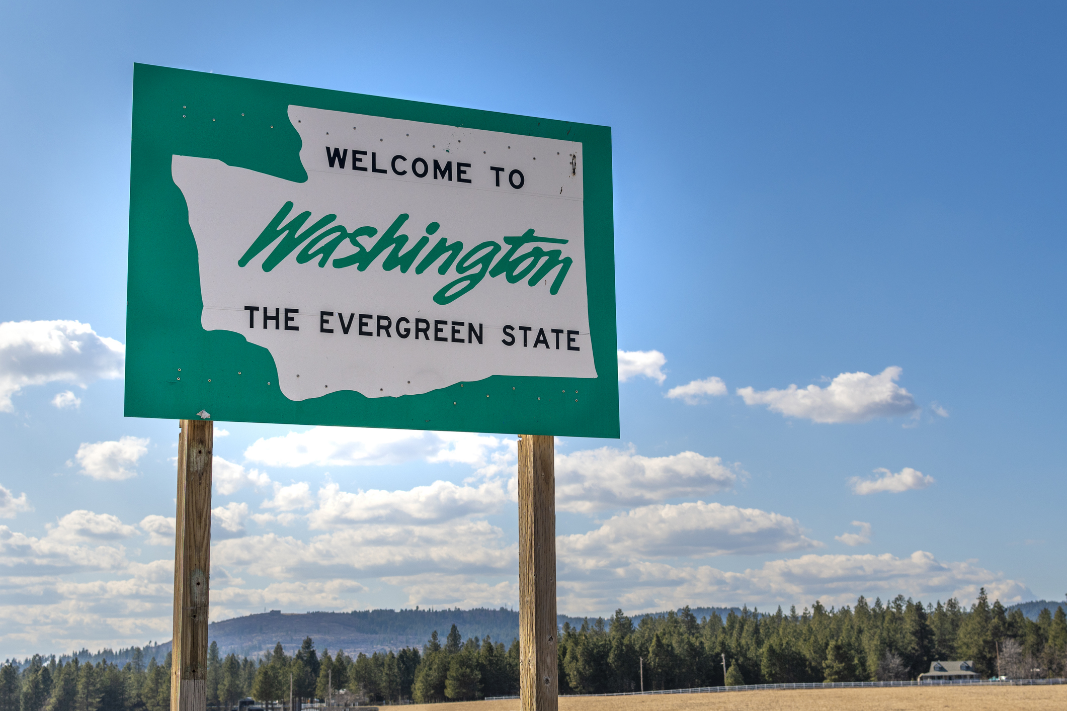A large roadside sign reads &quot;Welcome to Washington, The Evergreen State&quot; against a backdrop of a clear sky and forested landscape