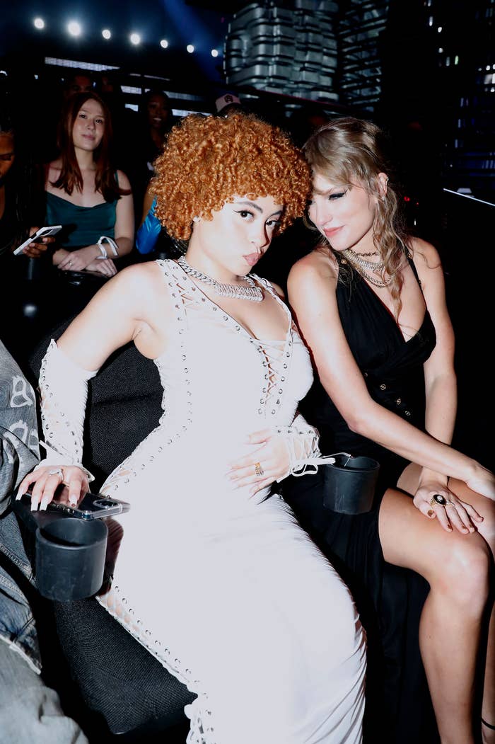 Ice Spice, in a dress with arm sleeves, sits beside Taylor Swift in a dress at an event. Both appear to be engaging with the camera