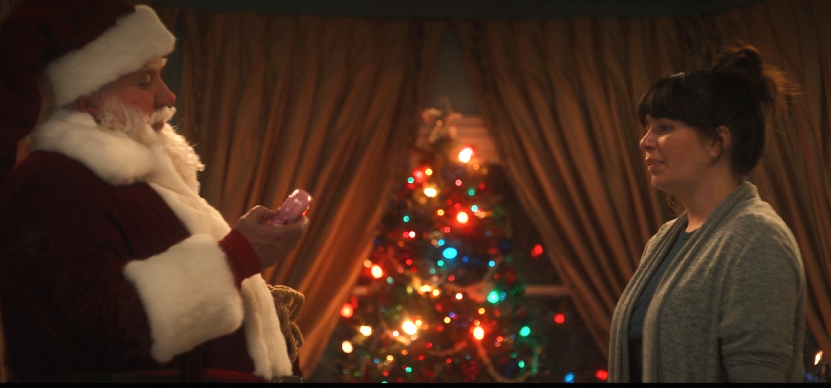 Santa Claus holds up a Polly Pocket toy while Casey&#x27;s character, in a cardigan, watches him. There is a decorated Christmas tree in the background