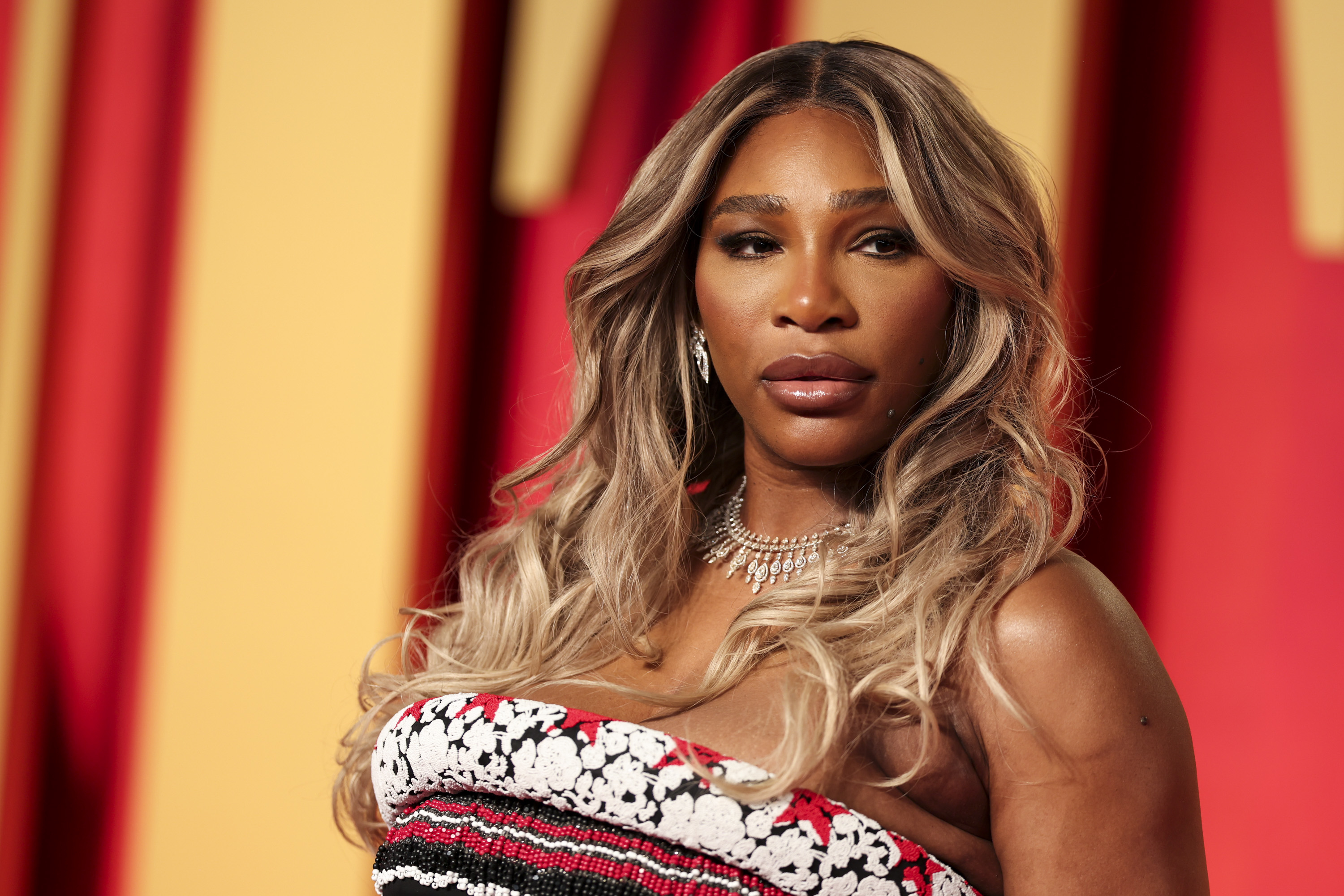 Serena Williams is wearing a strapless patterned dress and a diamond necklace on a red carpet