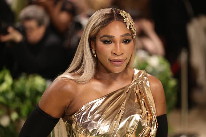 Serena Williams at a celebrity event, wearing a one-shoulder metallic gold dress and a floral-themed hair accessory