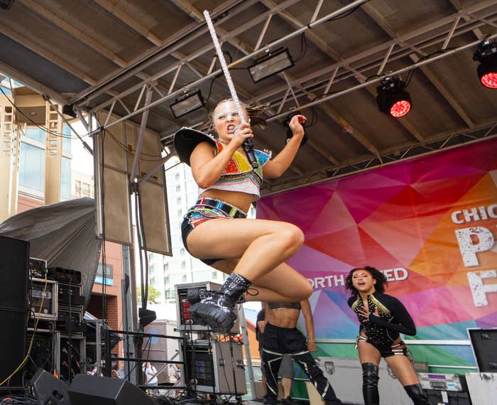 Performers on a festival stage, centered on a person mid-jump wearing a sparkling costume and boots, with backup dancers in the background