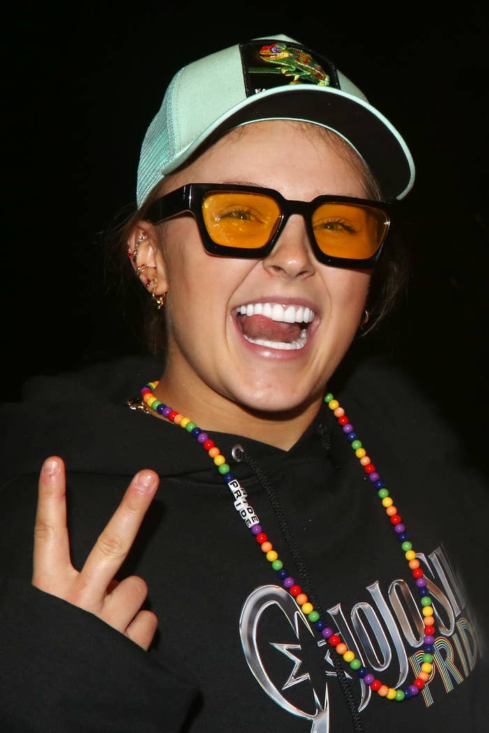JoJo Siwa smiling, wearing a cap, tinted glasses, beaded necklace, and hoodie, making a peace sign