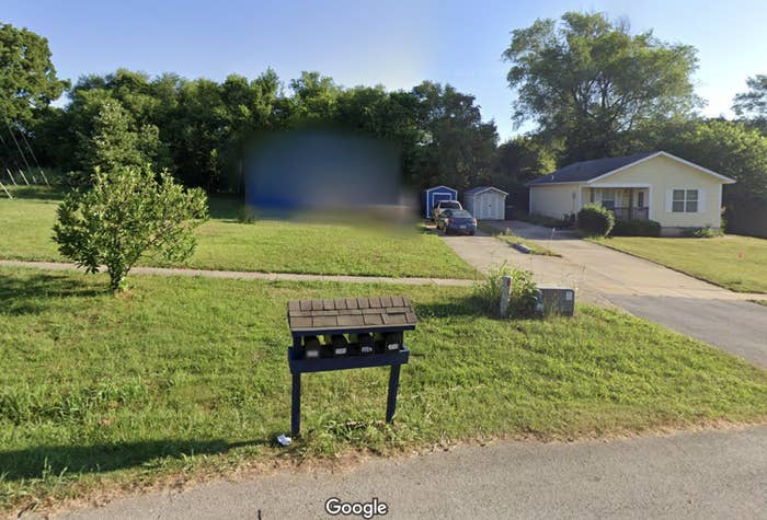 A Google Street View image showing a suburban residential area with a driveway, house, parked cars, mailbox, and trees