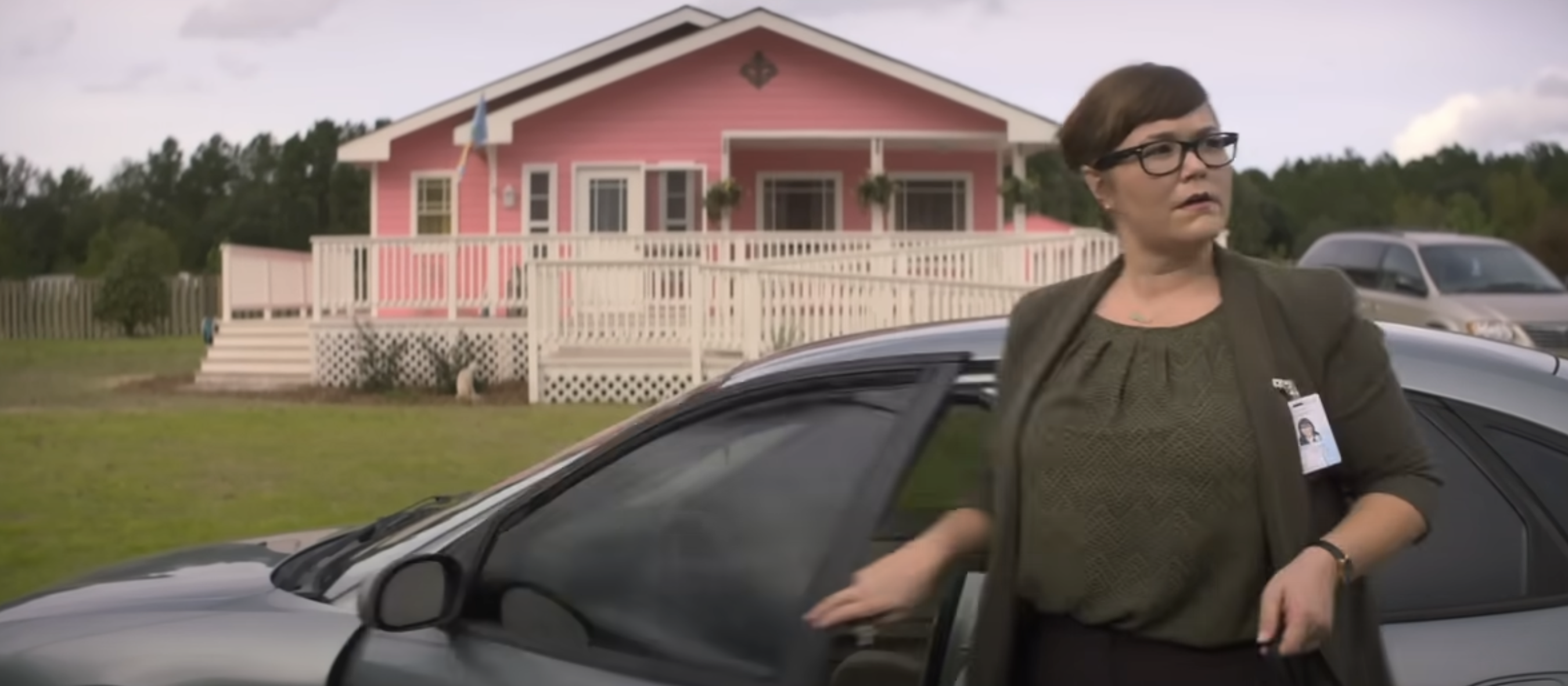 A woman wearing glasses and business attire with an ID badge steps out of a car in front of a house with a large porch