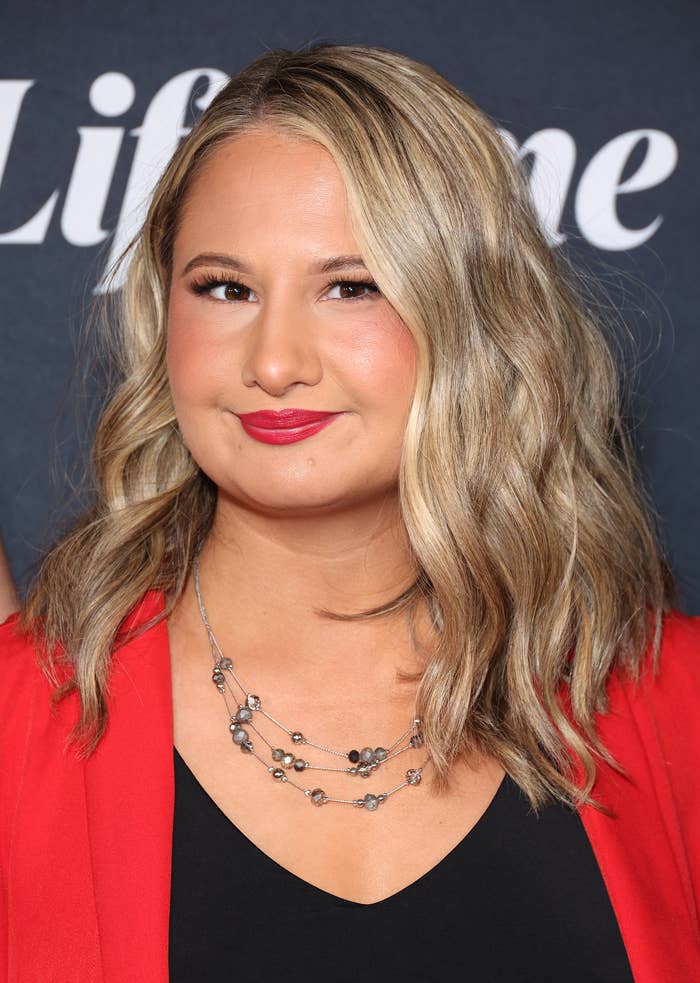 Woman with wavy hair in a black top, layered necklace, and red jacket at a Lifetime event