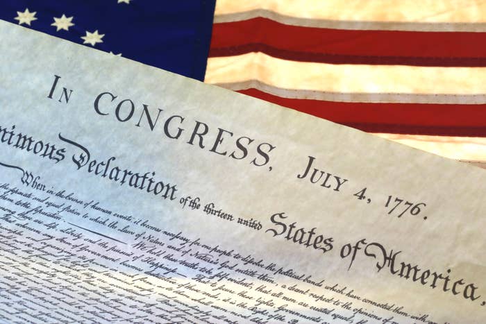 Declaration of Independence document dated July 4, 1776, with a partial view of an American flag in the background