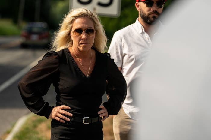 Marjorie Taylor Greene stands outdoors, wearing sunglasses and a black blouse with puffed sleeves. A man in a white shirt and red cap stands behind her