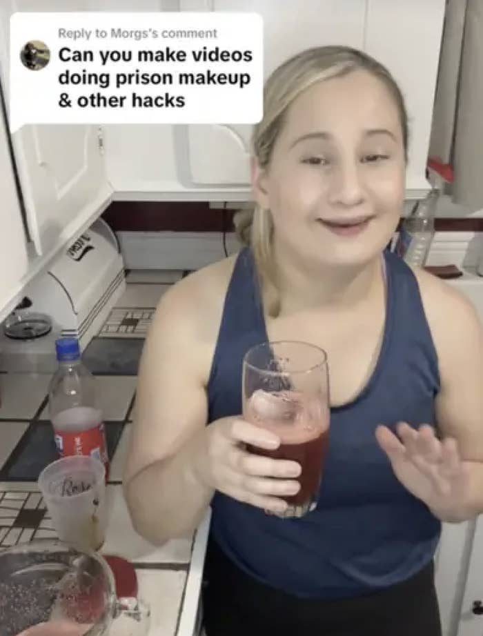 A woman in a kitchen holds a glass and smiles at the camera. A comment overlay asks, &quot;Can you make videos doing prison makeup &amp;amp; other hacks?&quot;