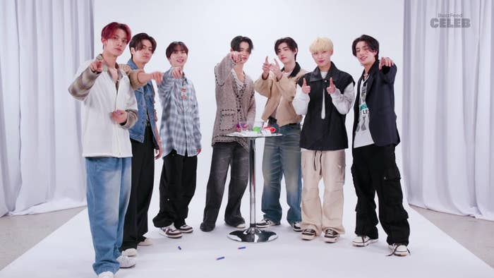 Members of Enhypen pose together pointing towards the camera. They are dressed in casual streetwear, including jeans, hoodies, and jackets