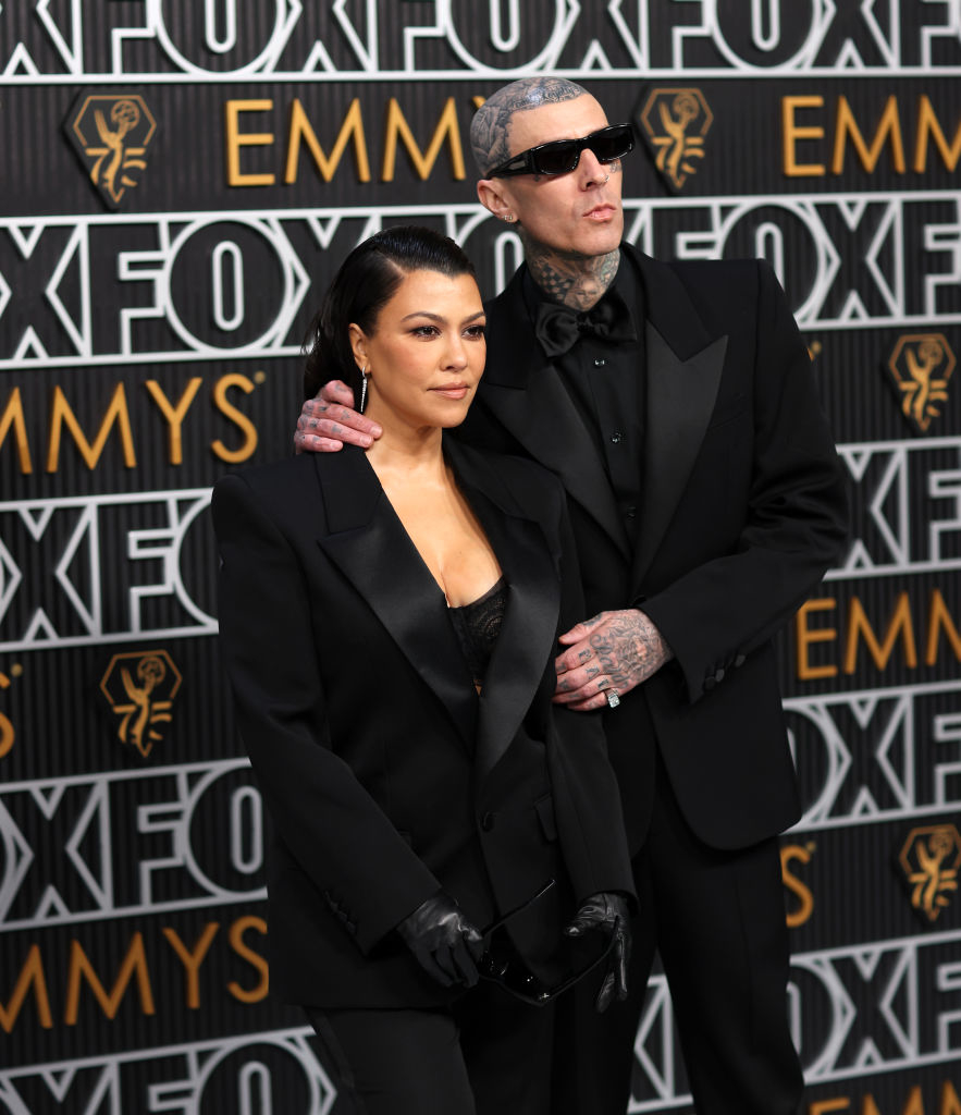Kourtney Kardashian and Travis Barker on a red carpet. Kourtney wears a tailored black suit, while Travis wears a black suit with sunglasses