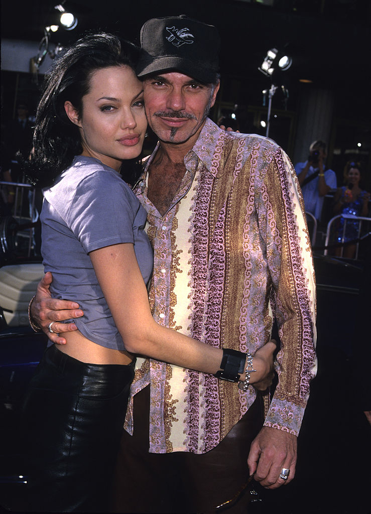 Angelina Jolie in a casual top and leather pants, hugging Billy Bob Thornton, wearing a patterned shirt and cap, both smiling at a public event
