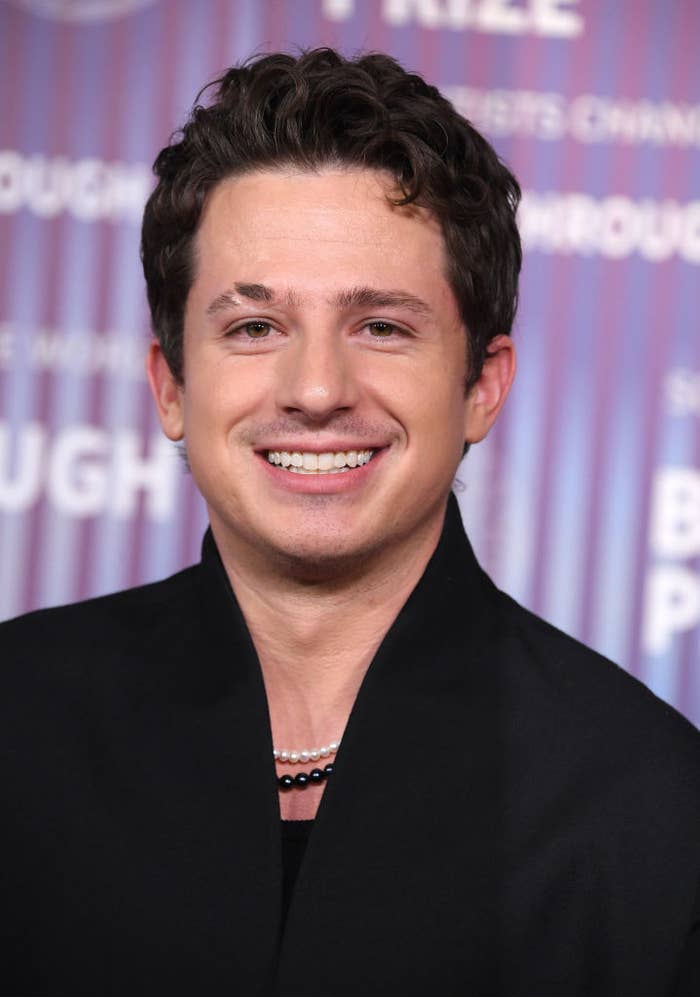 Charlie Puth smiles while wearing a black jacket and layered necklaces, standing in front of a striped background